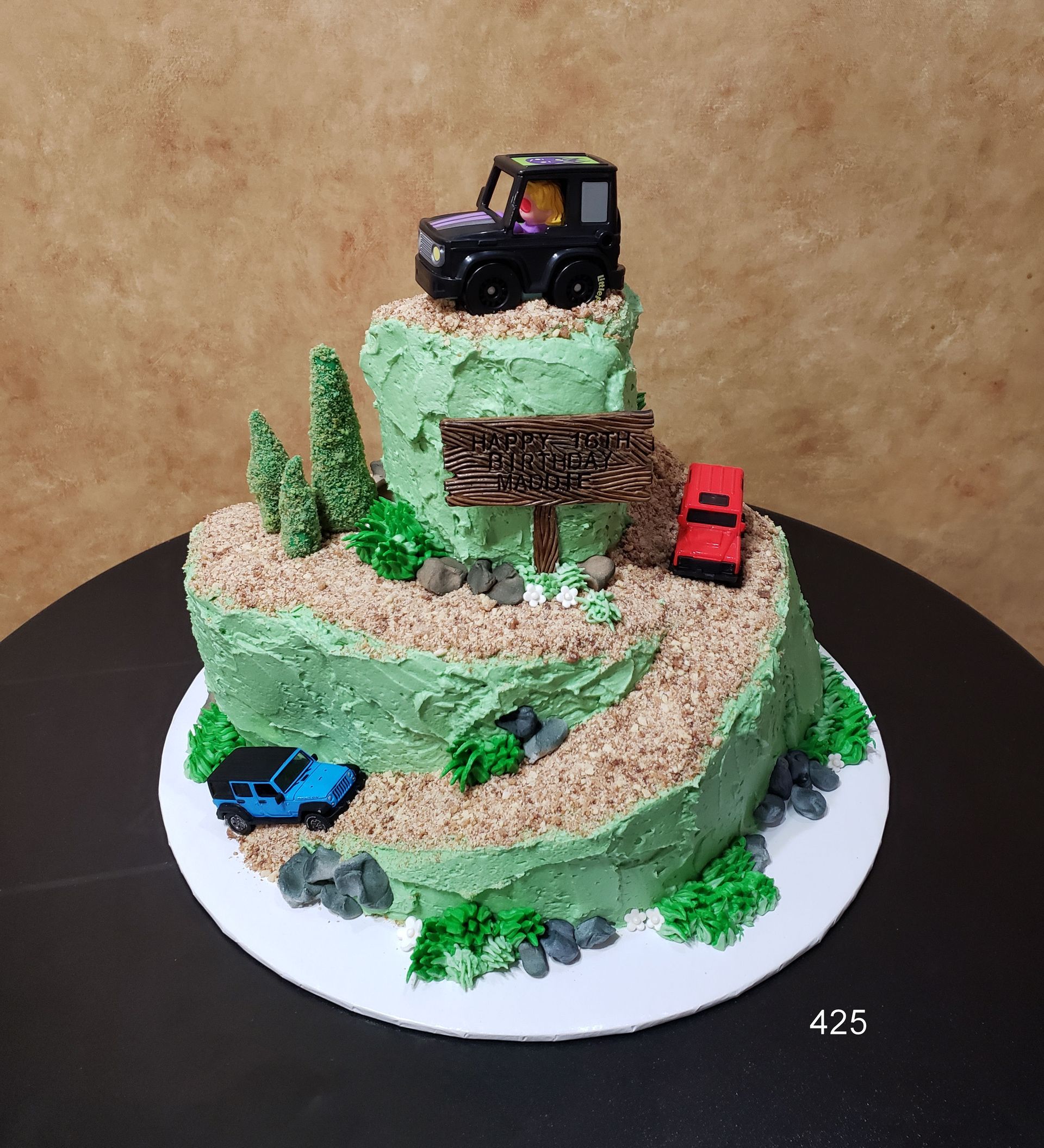 Tiered green cake diorama with toy trucks and trees on a white base