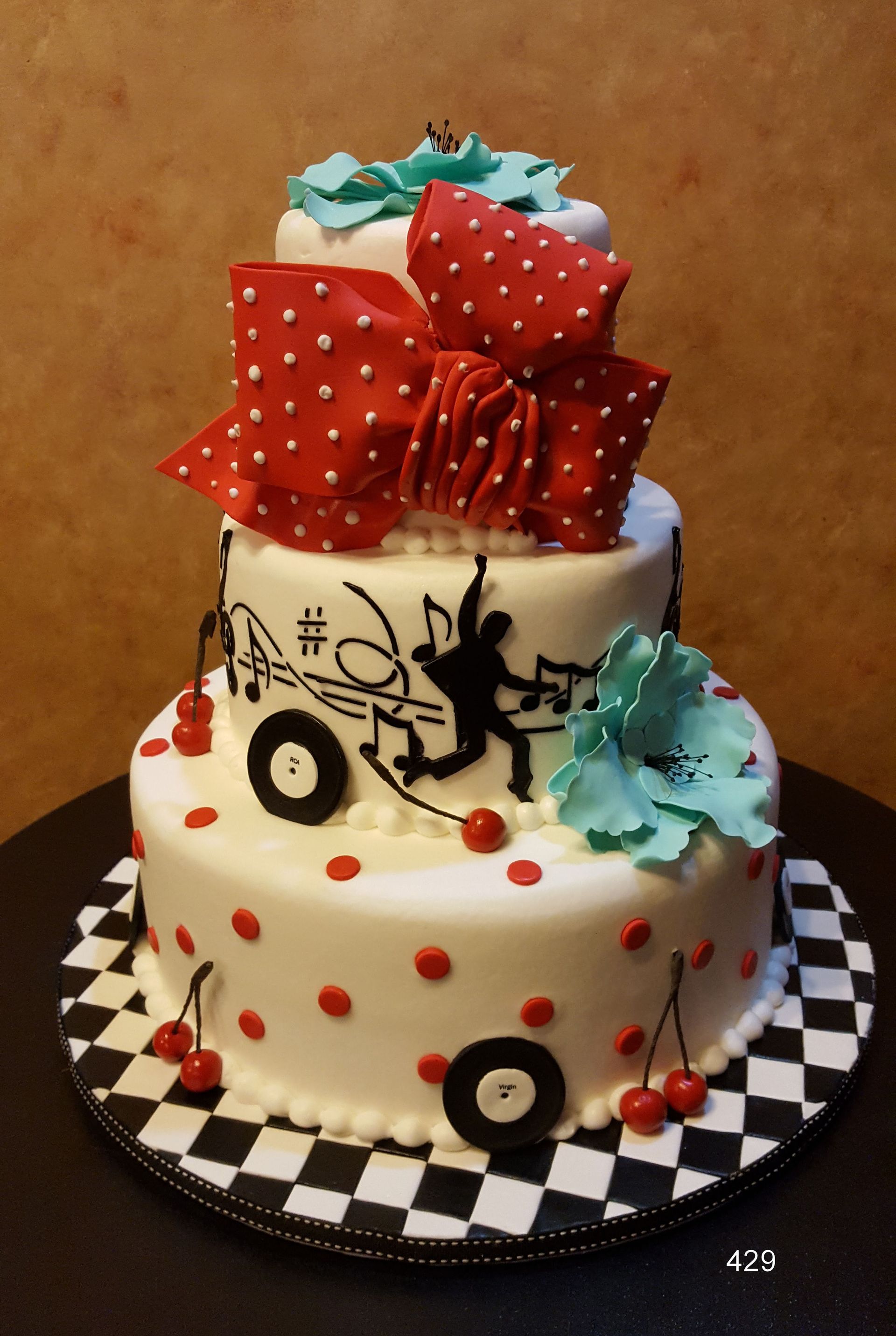 Two-tier cake with red polka-dot bow, teal frosting flowers, black-and-white checks, and red polka-dot accents.