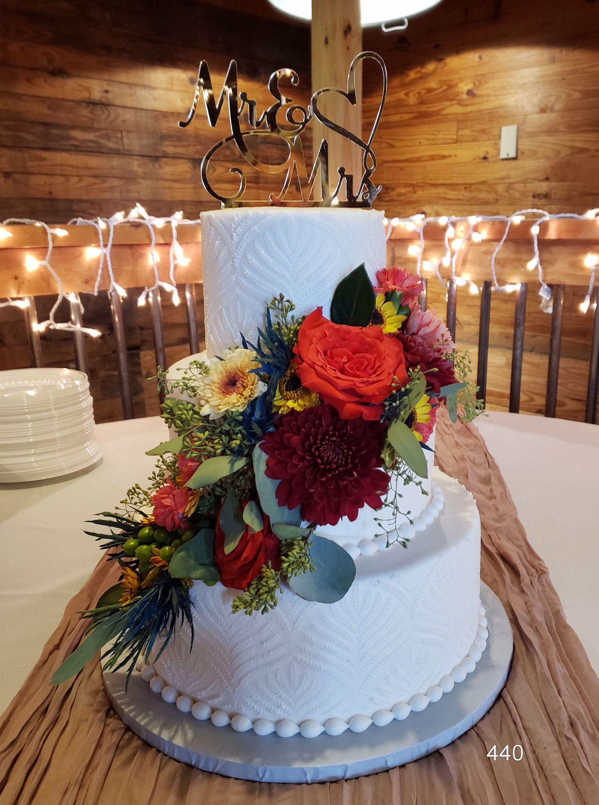 White wedding cake with colorful flowers on a decorated table in a warmly lit rustic venue