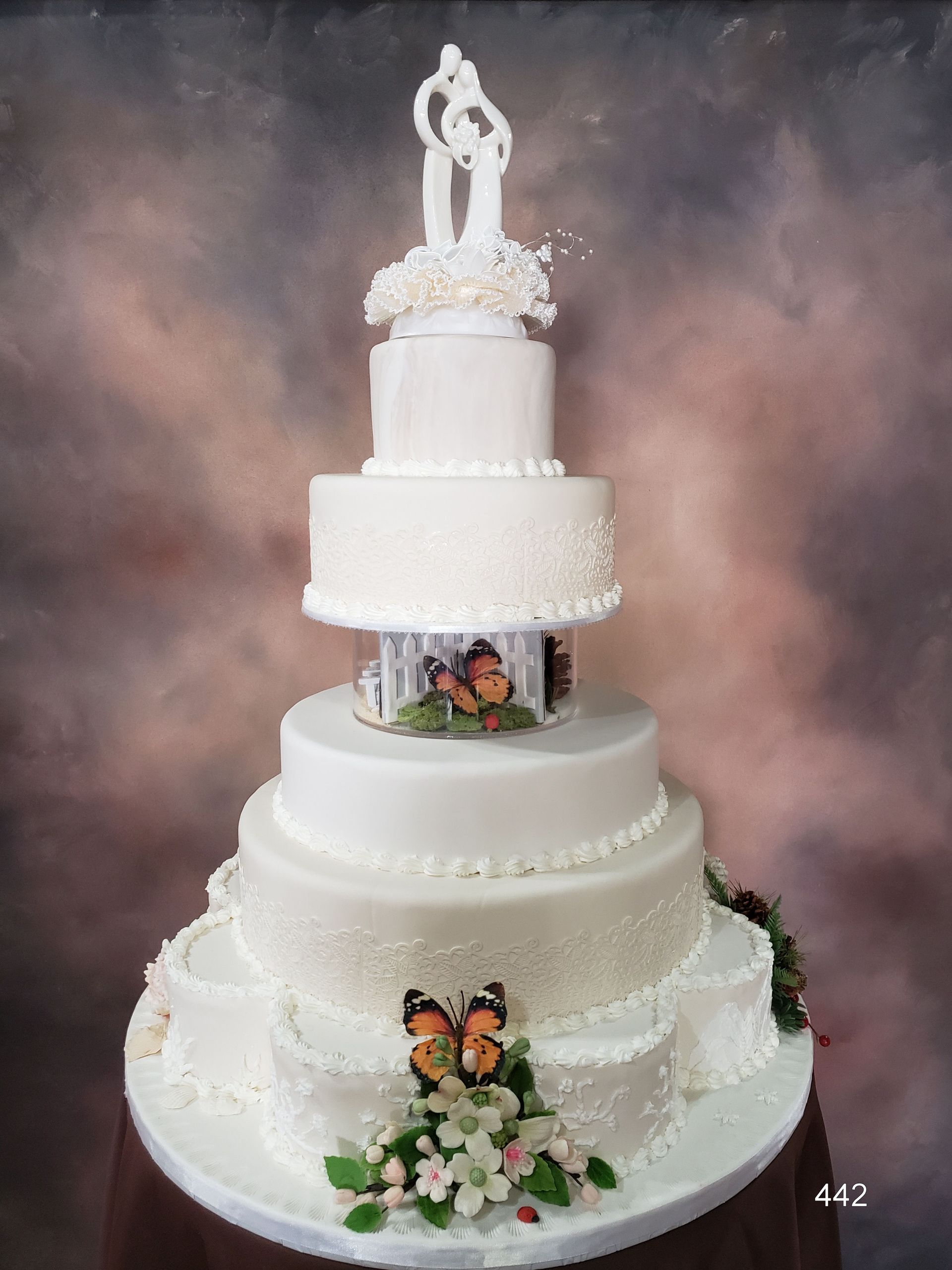 Five-tier white wedding cake with white flowers, butterflies, and a bride-and-groom topper on a smoky backdrop