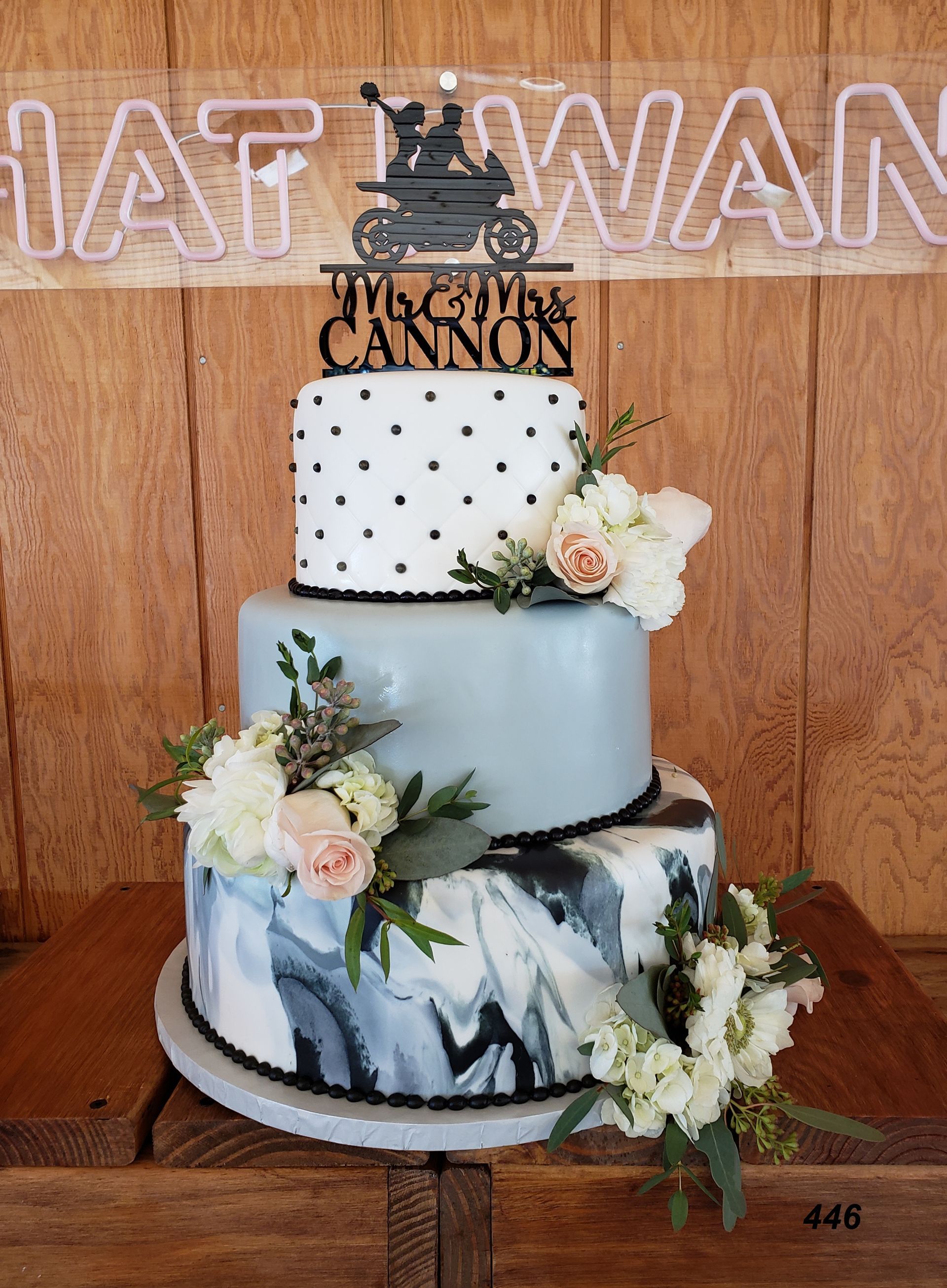 Three-tier wedding cake with blue, white, and floral accents on a wooden backdrop, topped with a “Mr. & Mrs.” sign