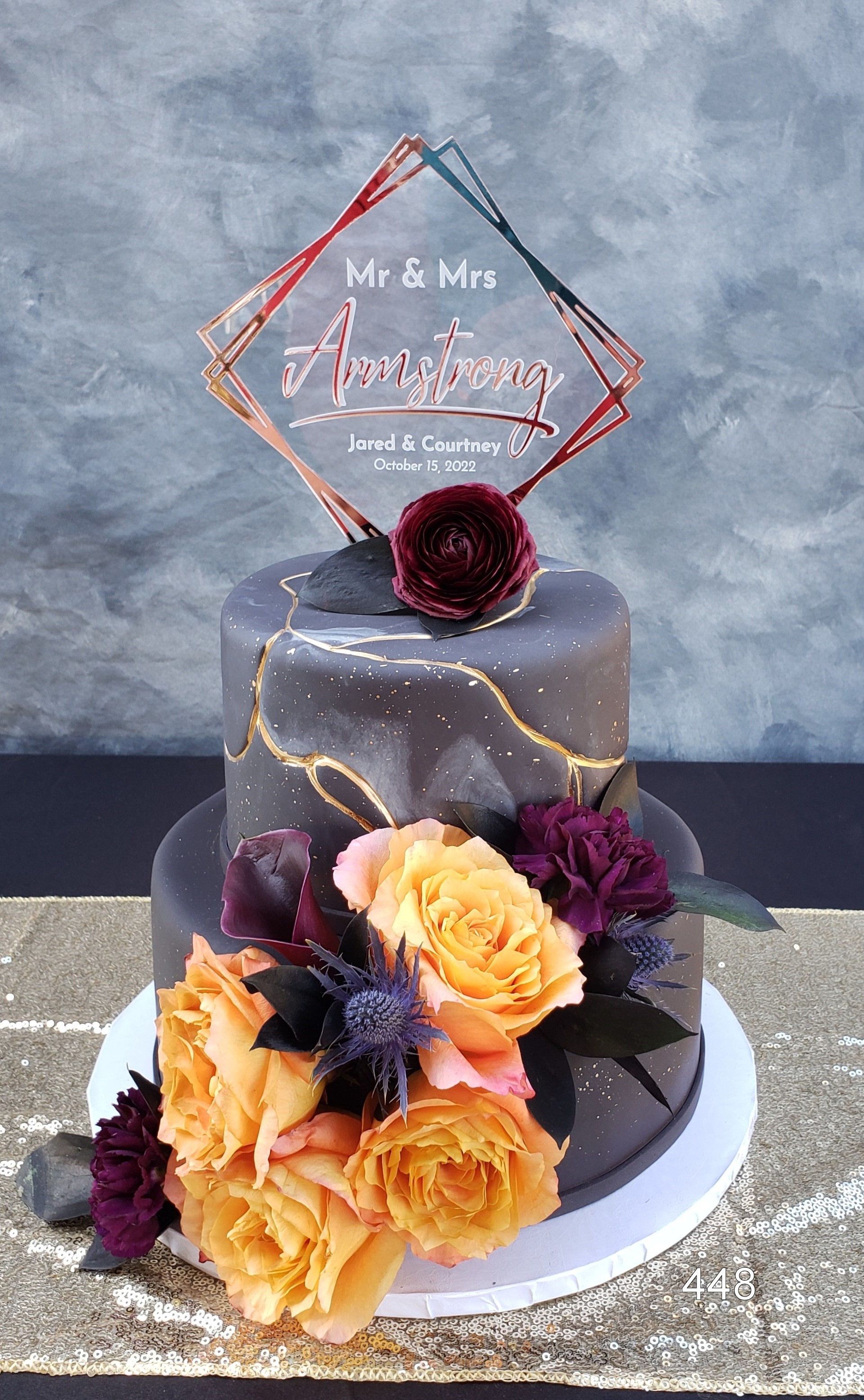 Decorative two-tier cake with gray icing, yellow and purple flowers, and a custom acrylic topper