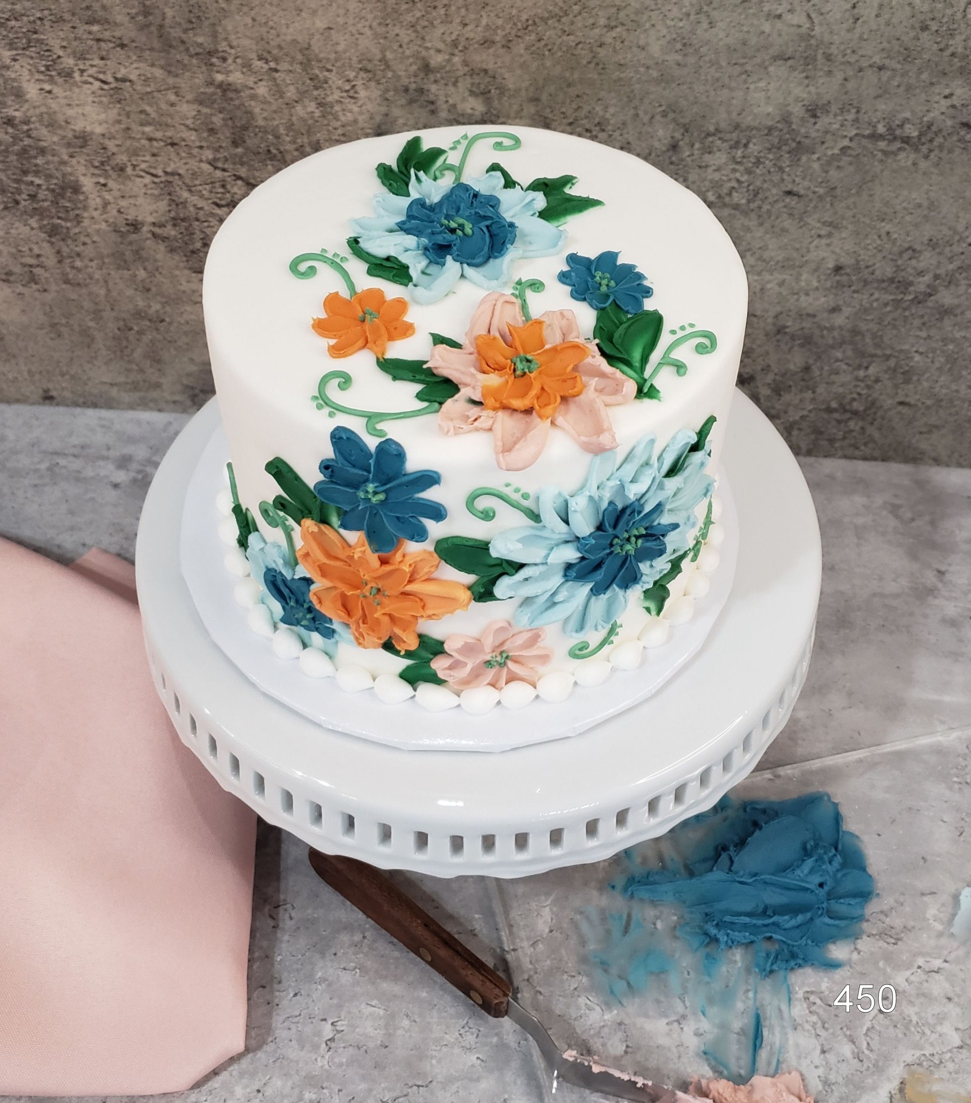 Small white cake with blue and orange floral icing on a turntable on a countertop