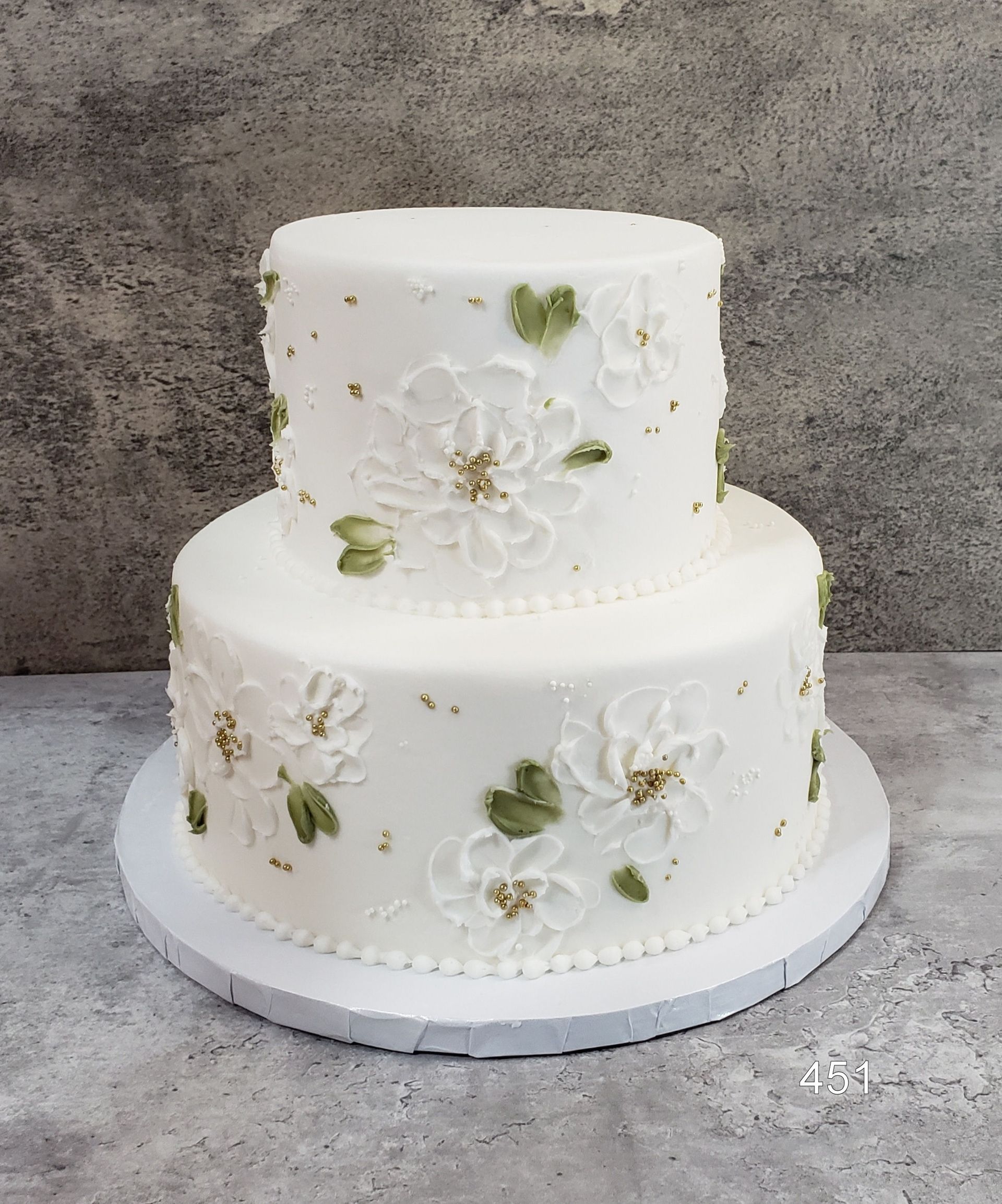 Two-tier white cake with green leaf accents and white floral decorations on a round board