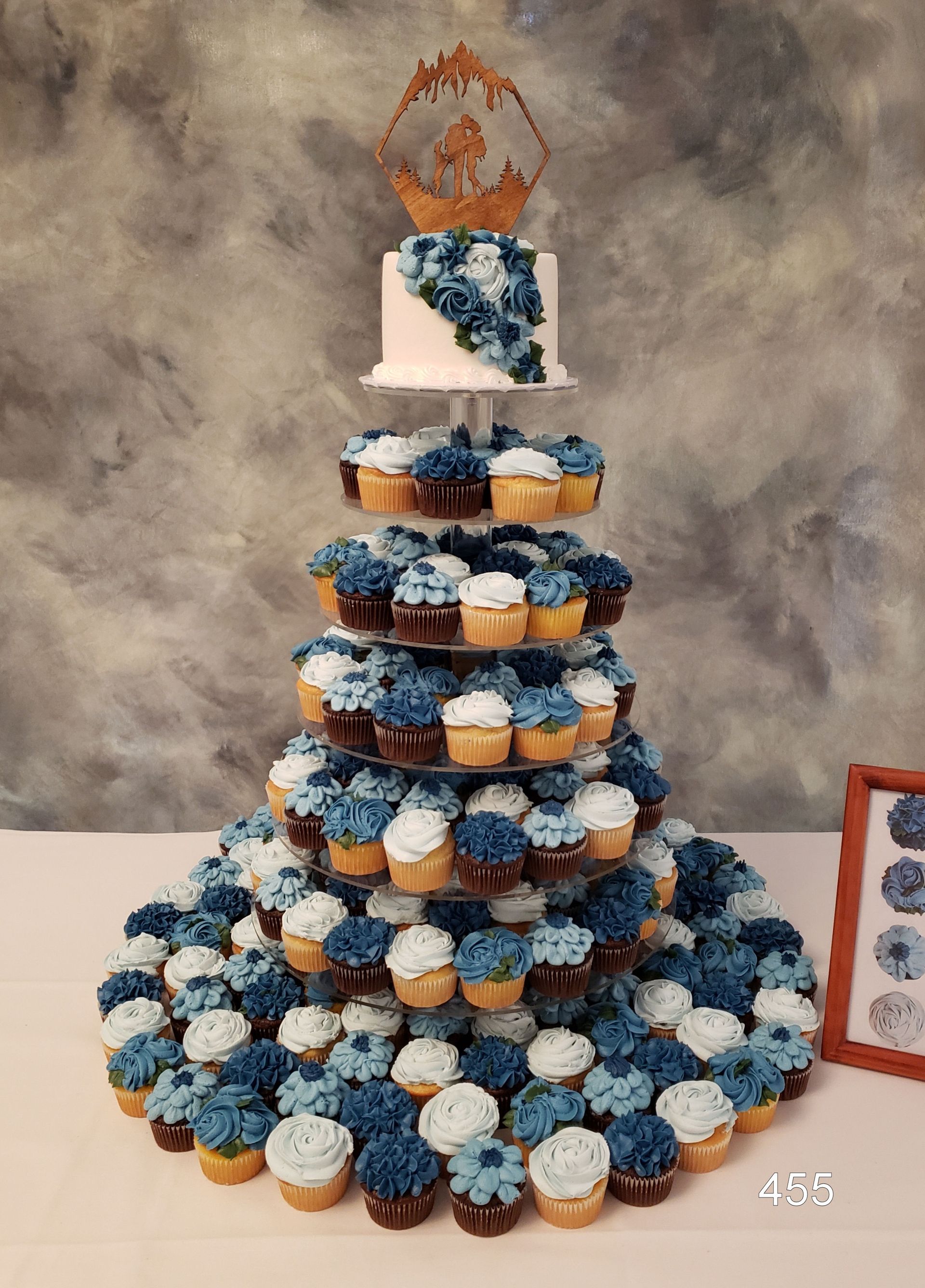 Tiered cupcake tower with blue, white, and peach cupcakes, topped by a small cake and decorative topper