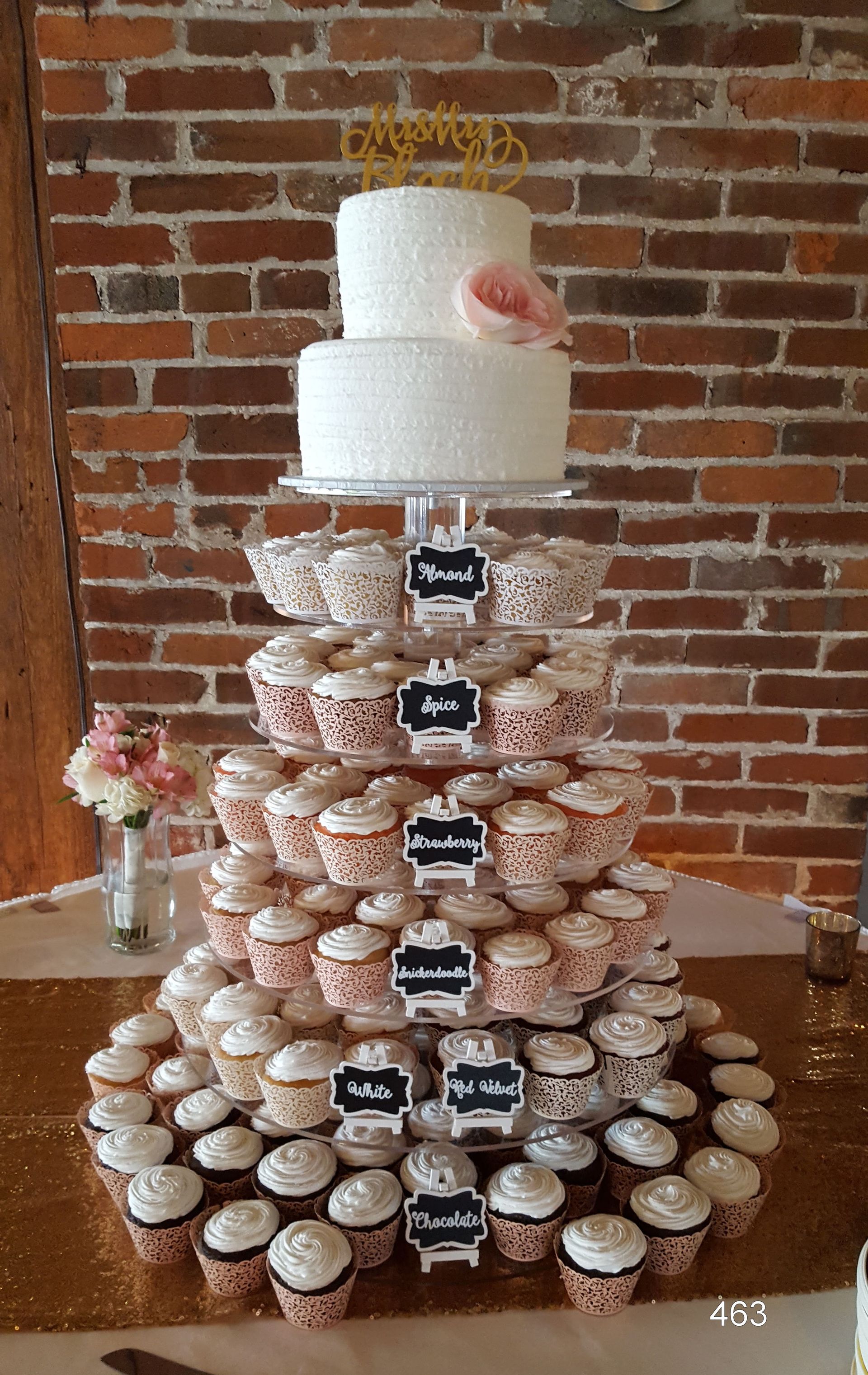 Tall tiered cake made of stacked cupcakes, topped with a white cake, on a table against a brick wall.