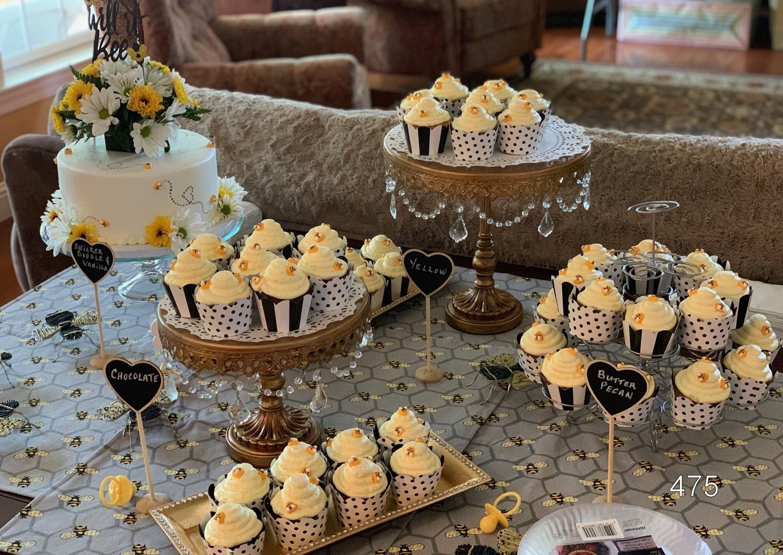 Dessert table with cupcakes and a white tiered cake on decorative stands in a living room.