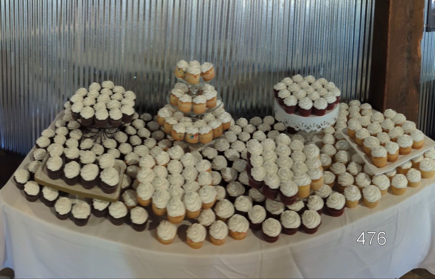 Dessert table with tiers of cupcakes and pastries on a white cloth against a corrugated metal backdrop