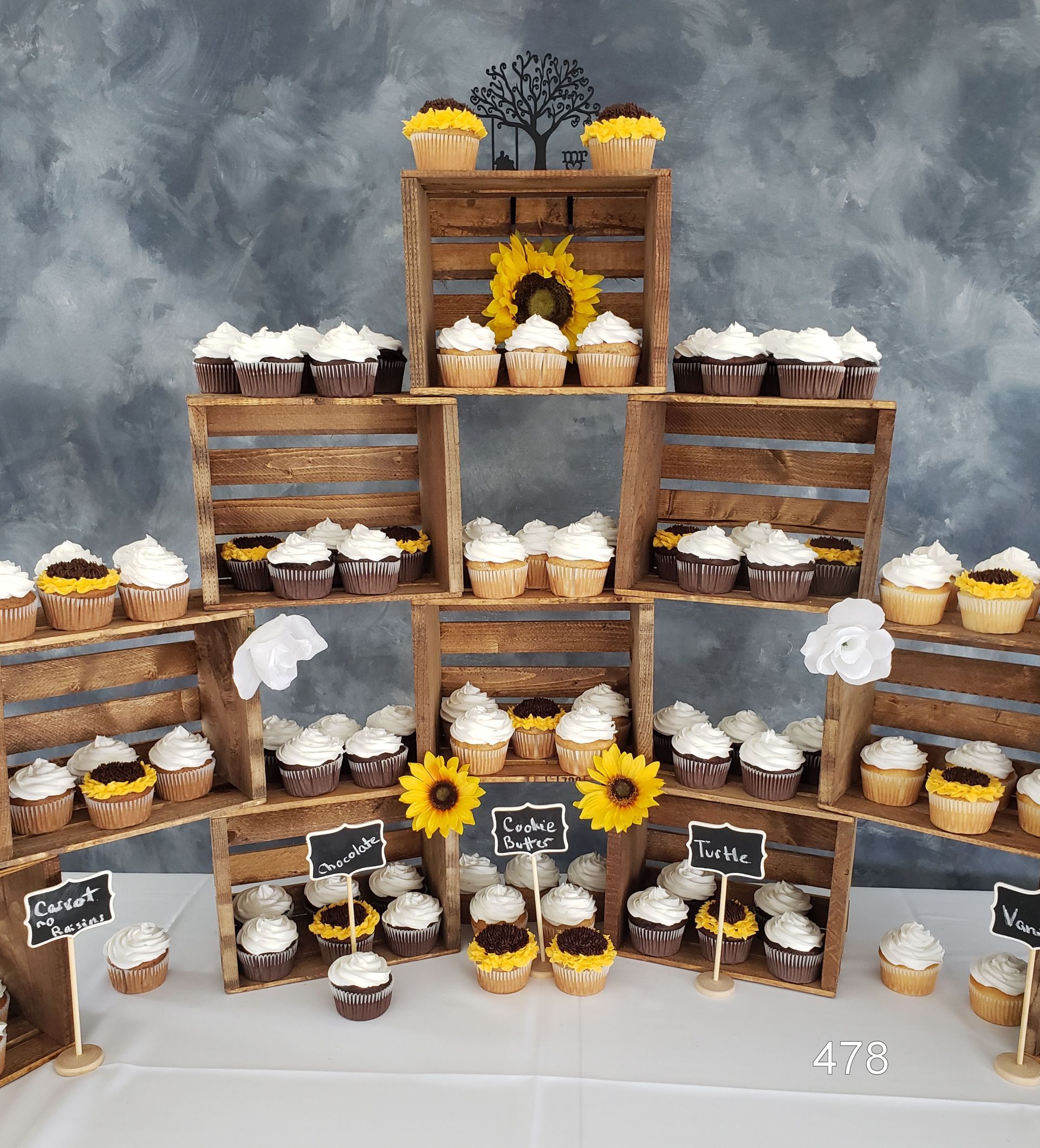 Tiered cupcake display with chocolate and white-frosted cupcakes, sunflowers, and rustic wooden crates on a gray backdrop