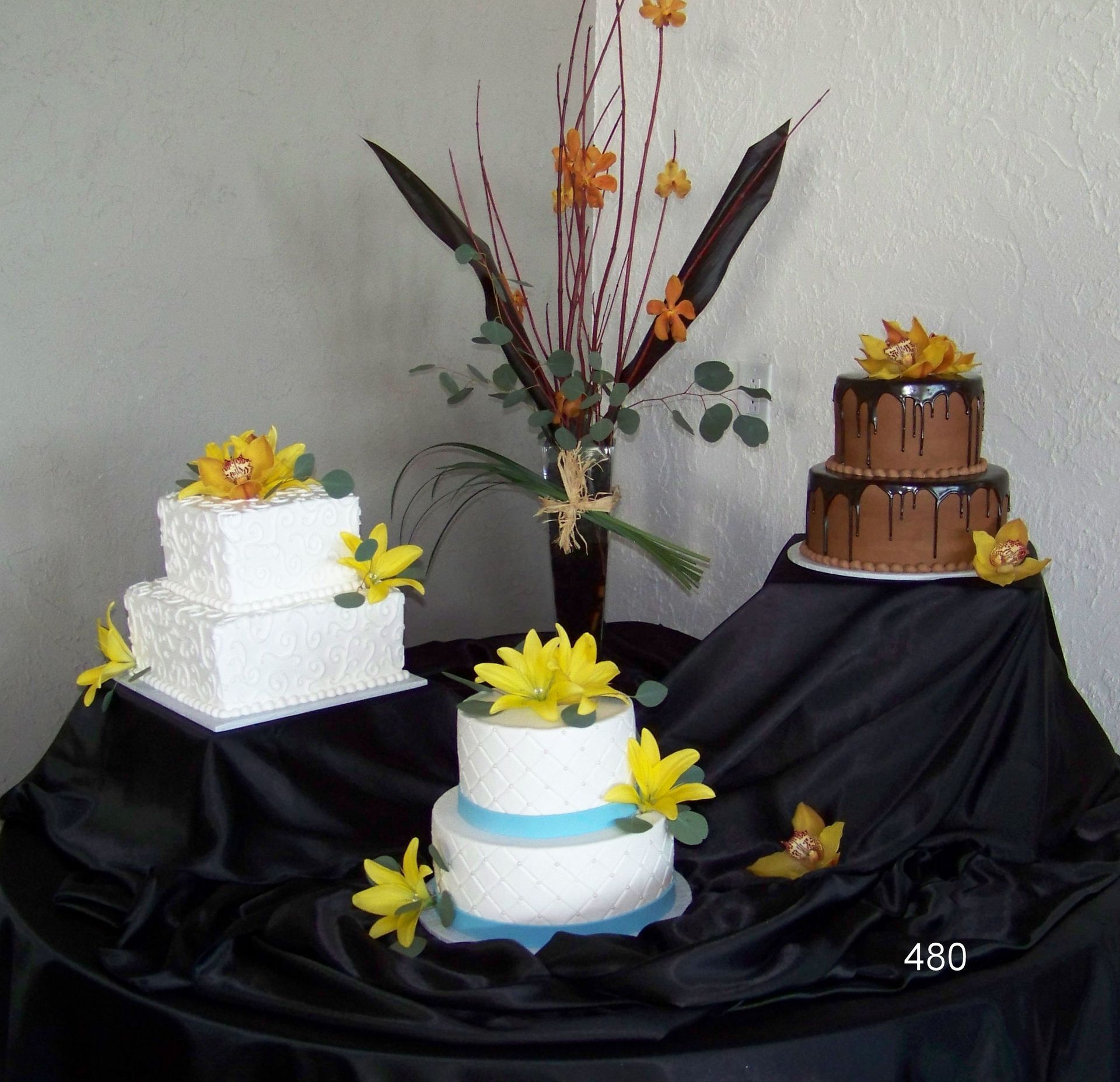 Assorted decorated cakes on black cloth with yellow flowers and a floral centerpiece on a table.