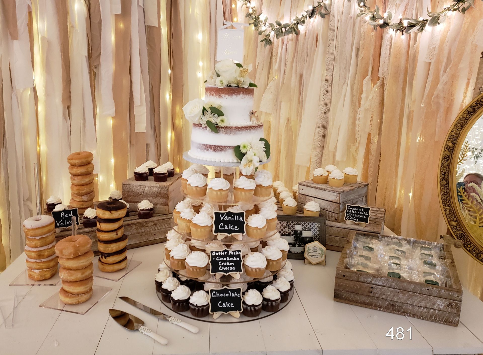 Elegant dessert table with tiered white cake, cupcakes, cookies, and pastries against a gold draped backdrop