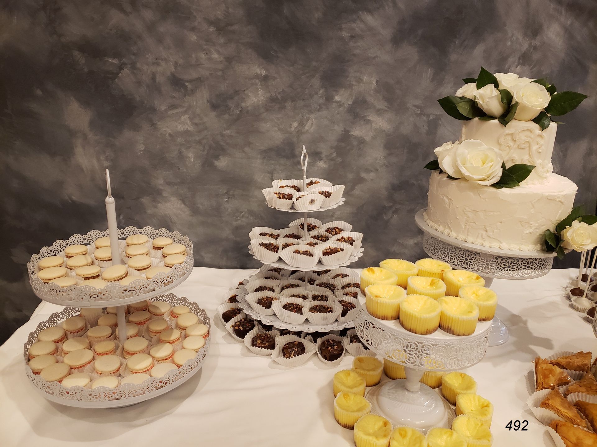 Assorted dessert trays and a tiered cake with white flowers on a table against a gray backdrop