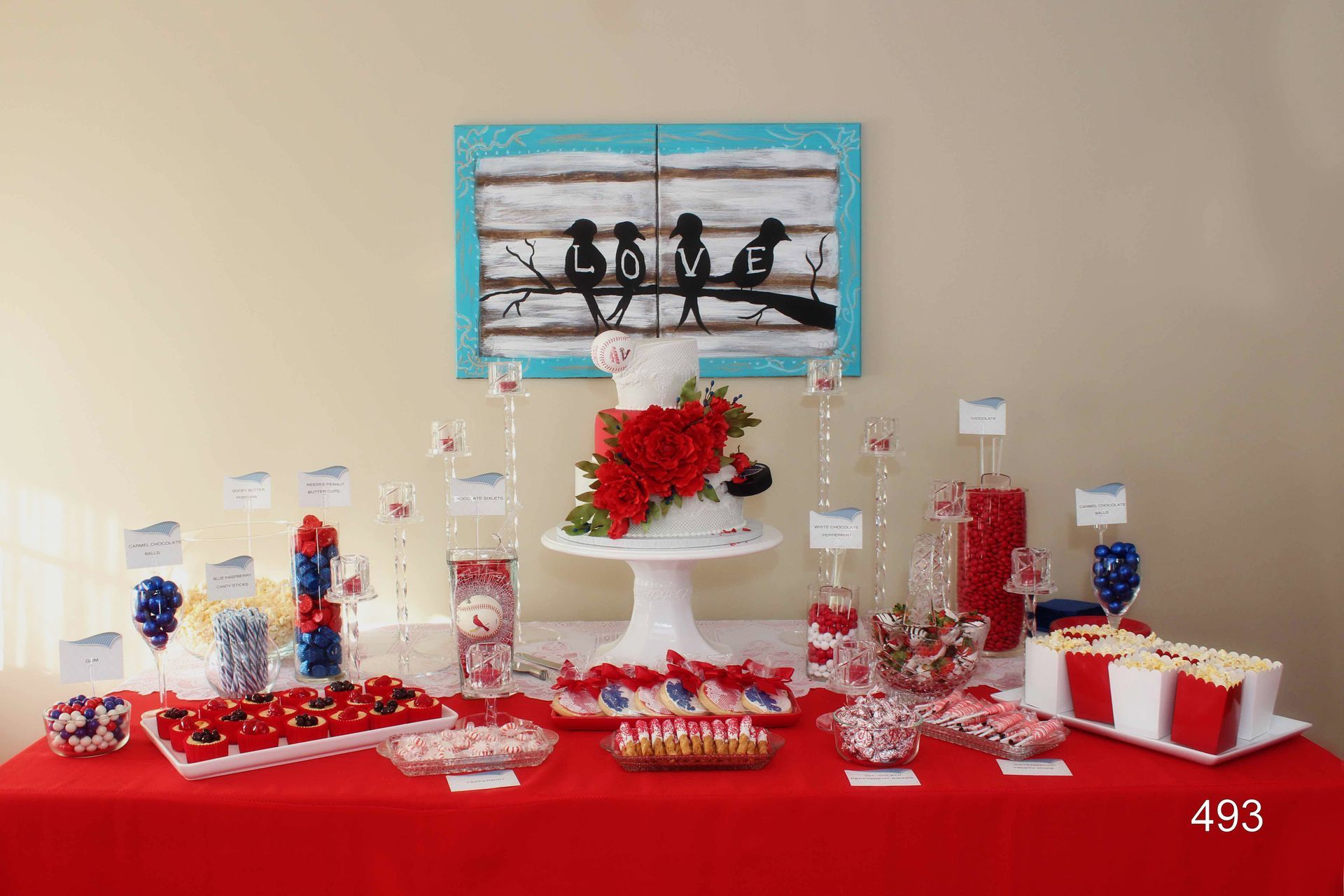 Red table with a dessert display, white cake stand, and blue framed artwork on a beige wall.