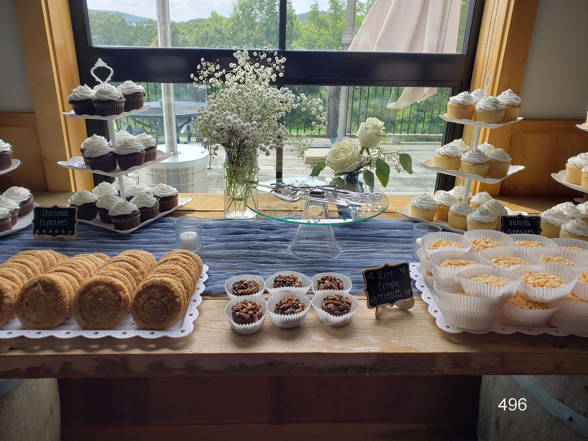 Dessert table with assorted pastries and cupcakes arranged on trays by a window with flowers in the center