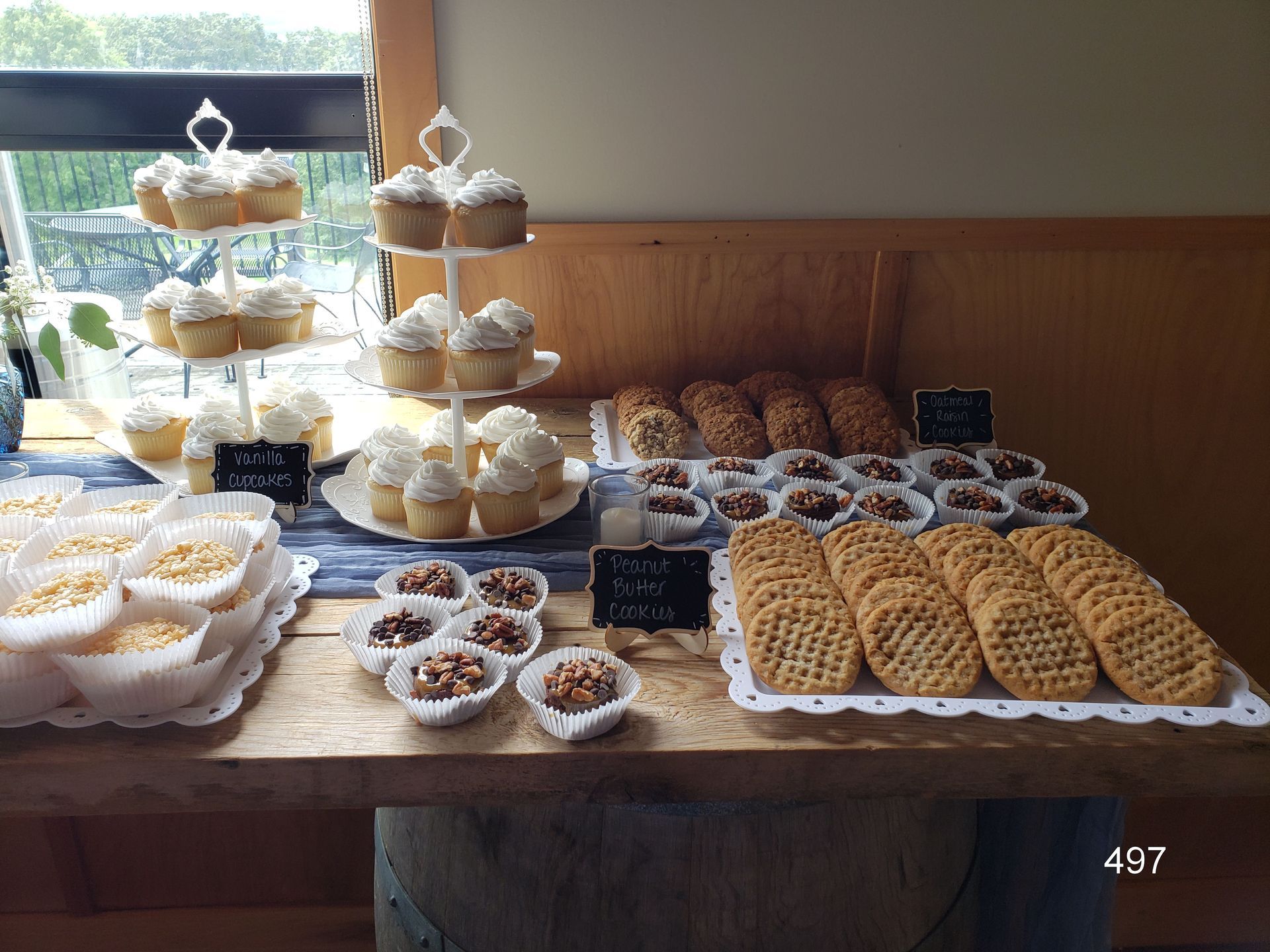 Dessert buffet with cupcakes, cookies, pastries, and sweets arranged on a wooden table by a window.
