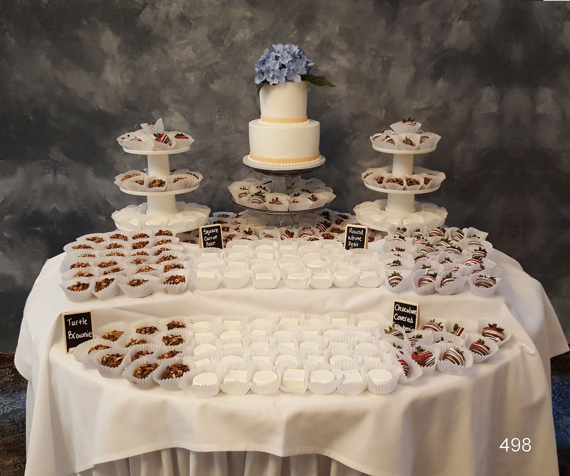White dessert table with a tiered blue-and-white cake, cupcake stands, and assorted sweets on a draped table