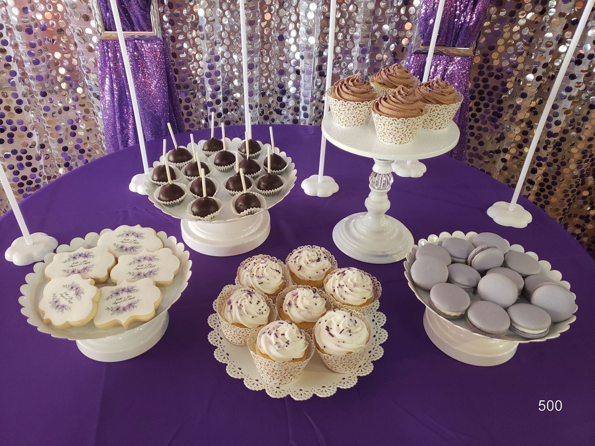 Dessert table with assorted cupcakes and cookies on white stands over a purple cloth backdrop