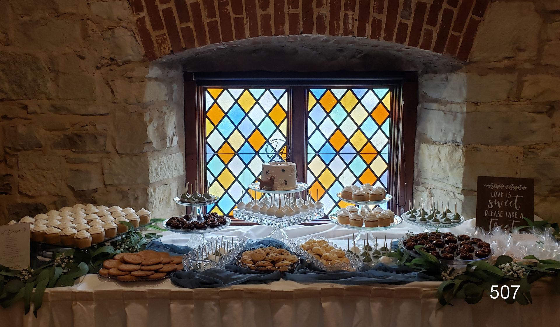 Rustic buffet table under stained-glass windows, with cakes, pastries, and candles on a stone wall backdrop