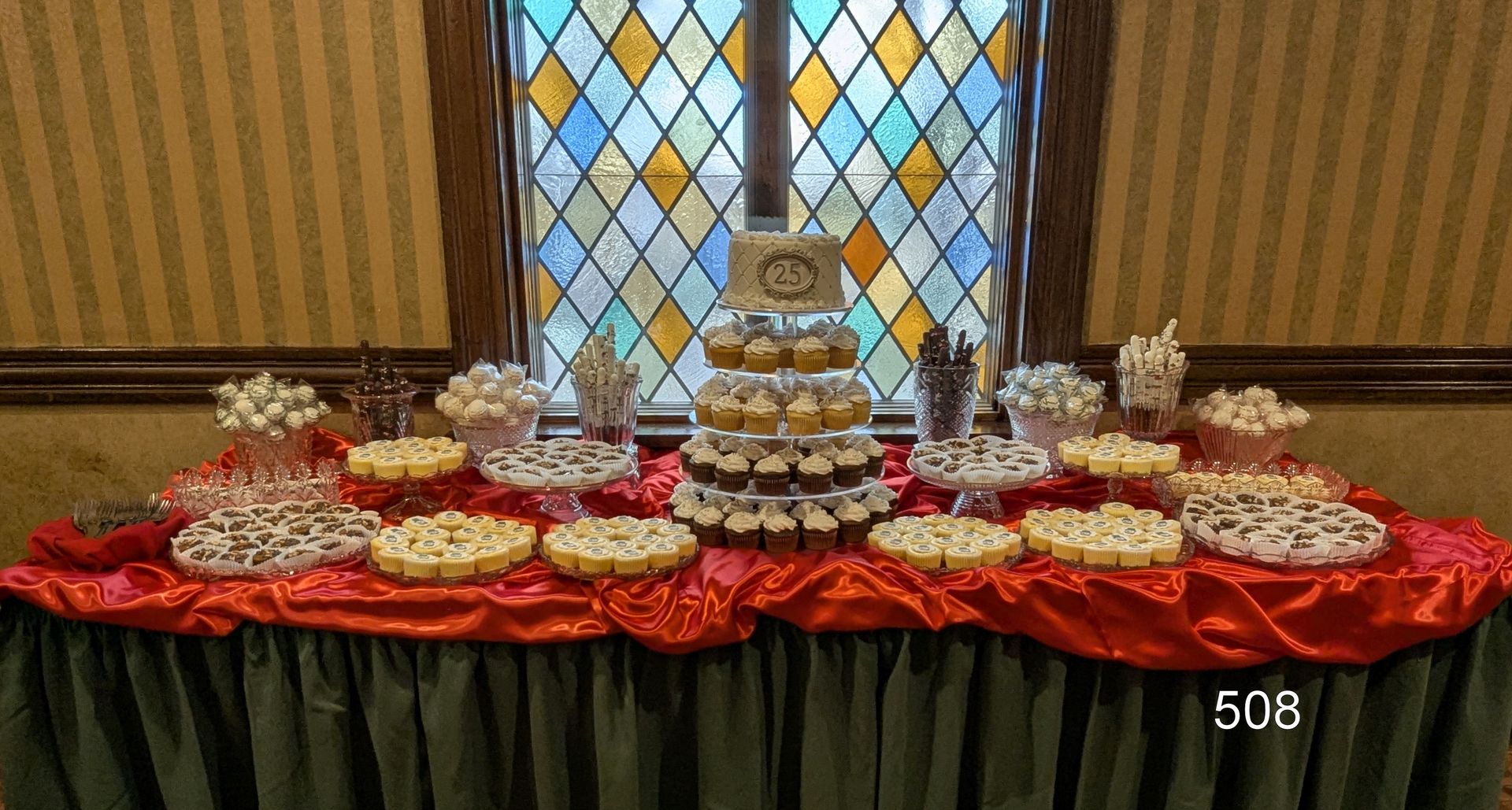 Table with assorted pastries and desserts on red cloth, arranged in tiers in front of a stained-glass window.