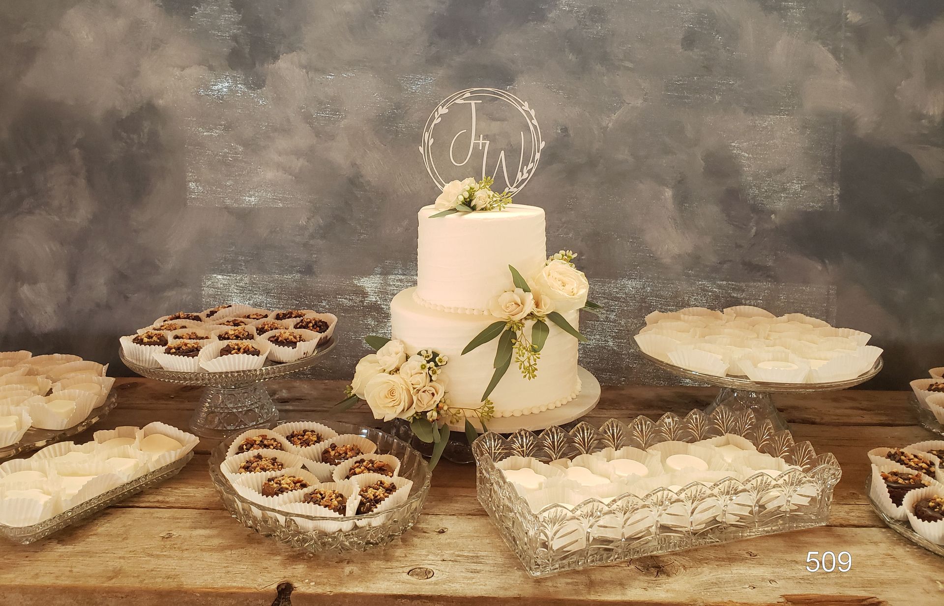White tiered cake with flowers on a rustic table, surrounded by assorted desserts at a dessert display.