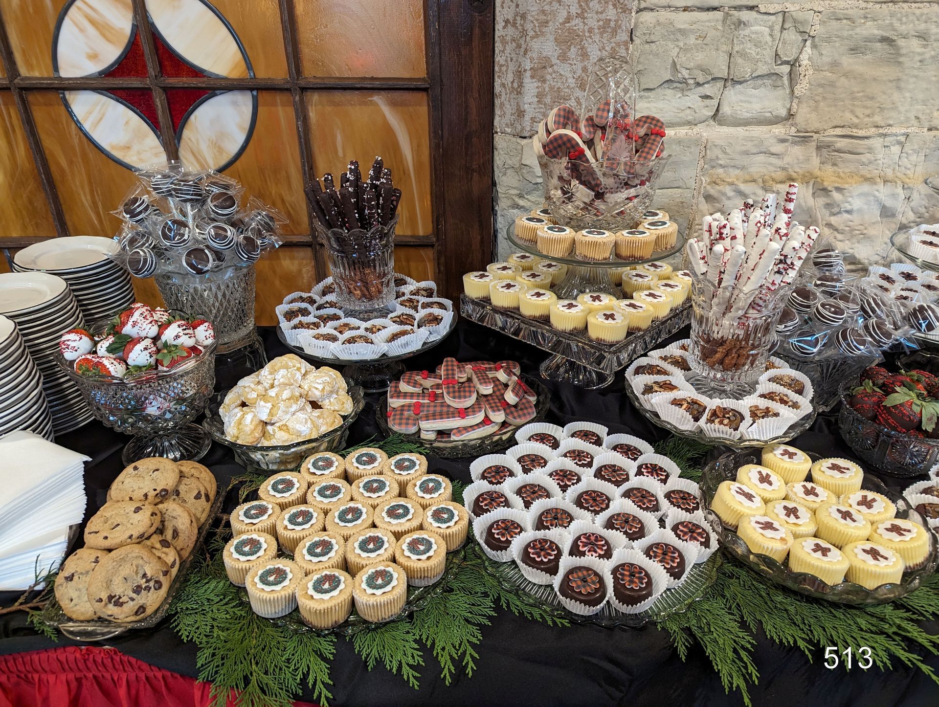 Assorted pastries and cookies arranged on a table with greenery, in front of a stone wall and stained glass window.