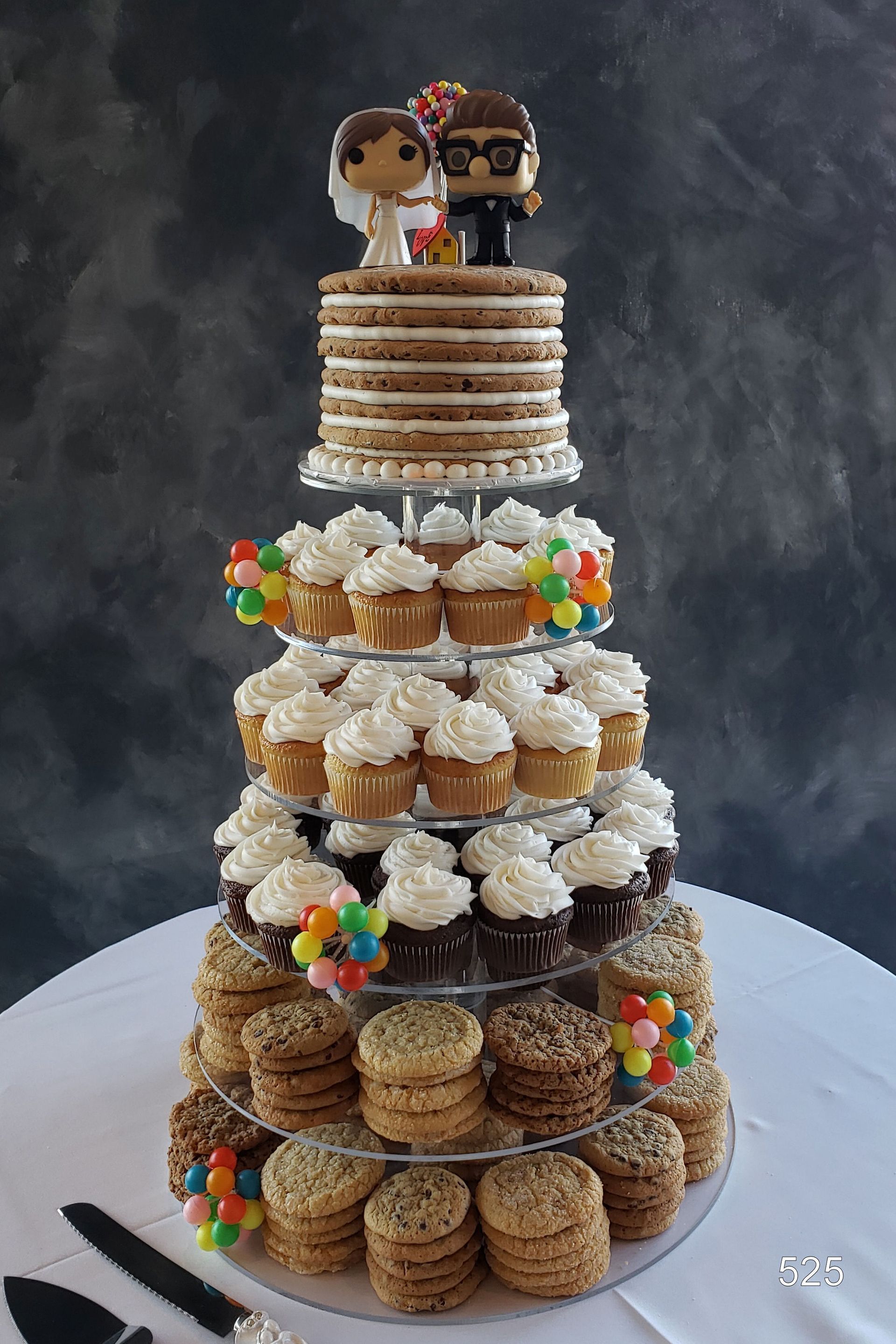Tiered dessert tower of stacked cupcakes and cookies topped with two figurines on a white table