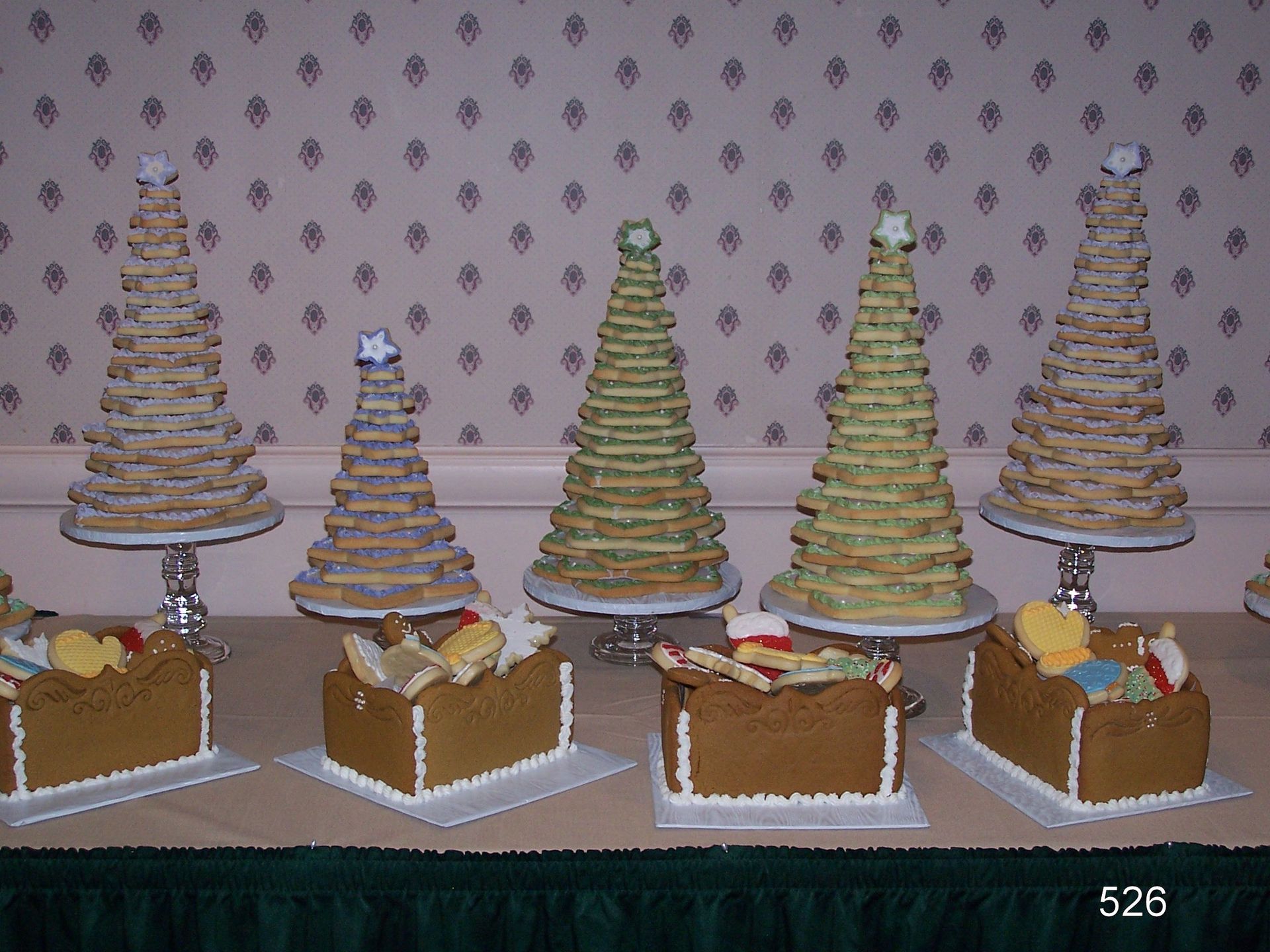 Rows of gingerbread houses topped with cone-shaped tree cookies on a table against patterned wallpaper.