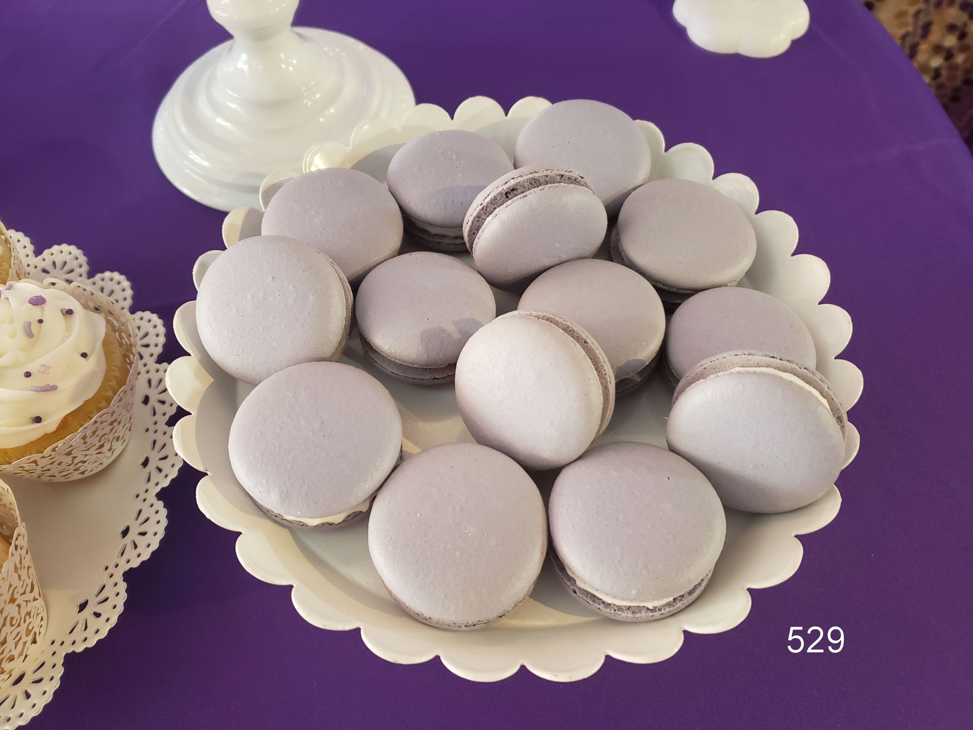Plate of lavender and white macarons on a purple tablecloth, with a white pedestal dish in the background