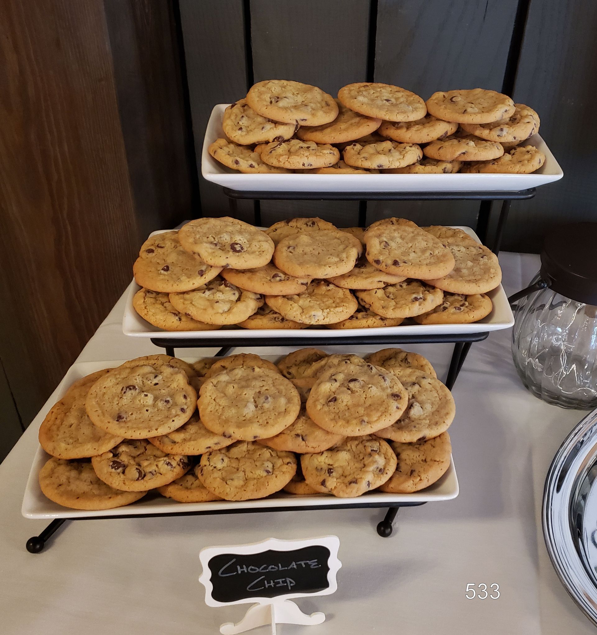 Three trays of chocolate chip cookies on a dessert table, with a small label in front.