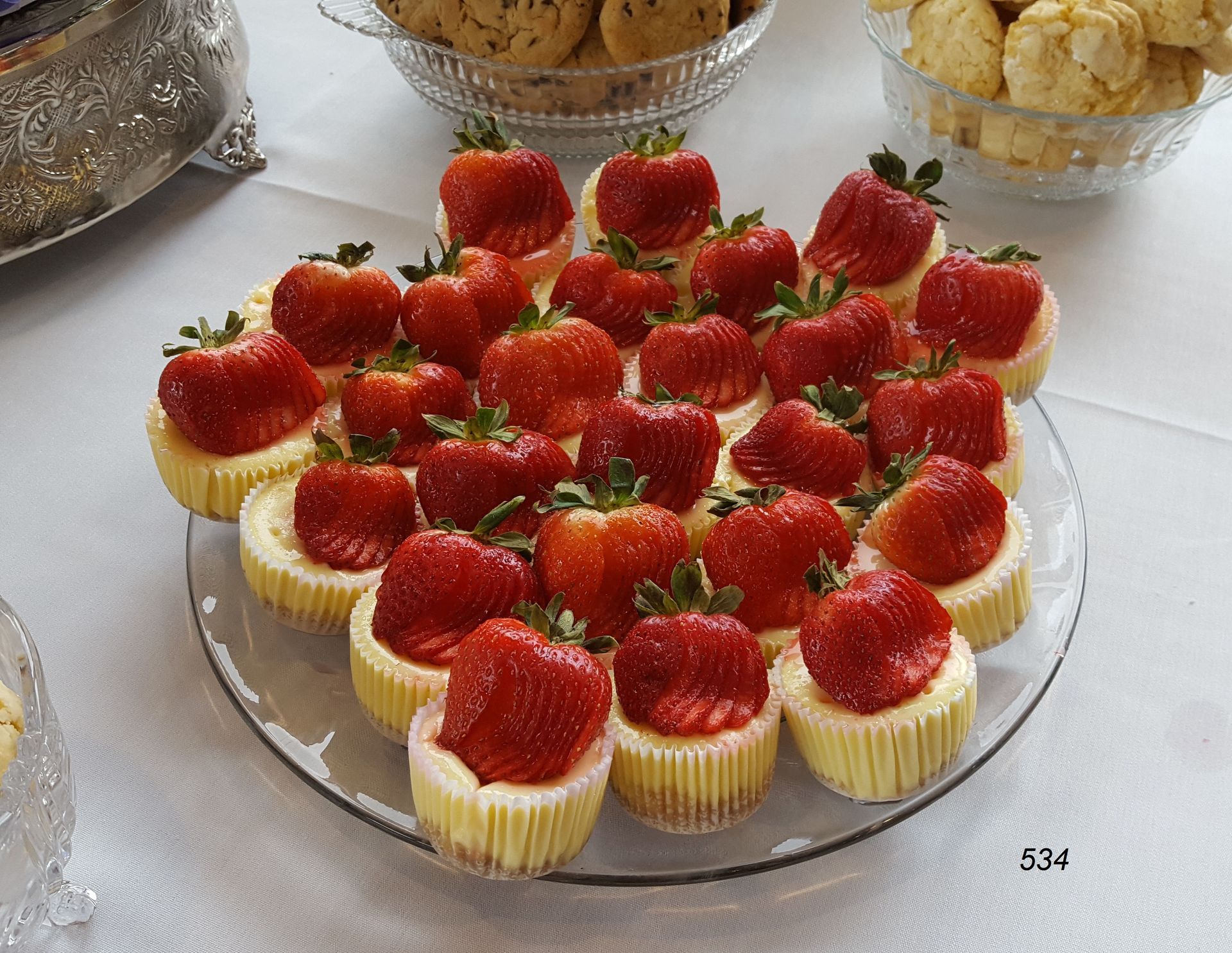 Tray of mini strawberry cupcakes on a table, topped with halved strawberries in a clear dessert platter.