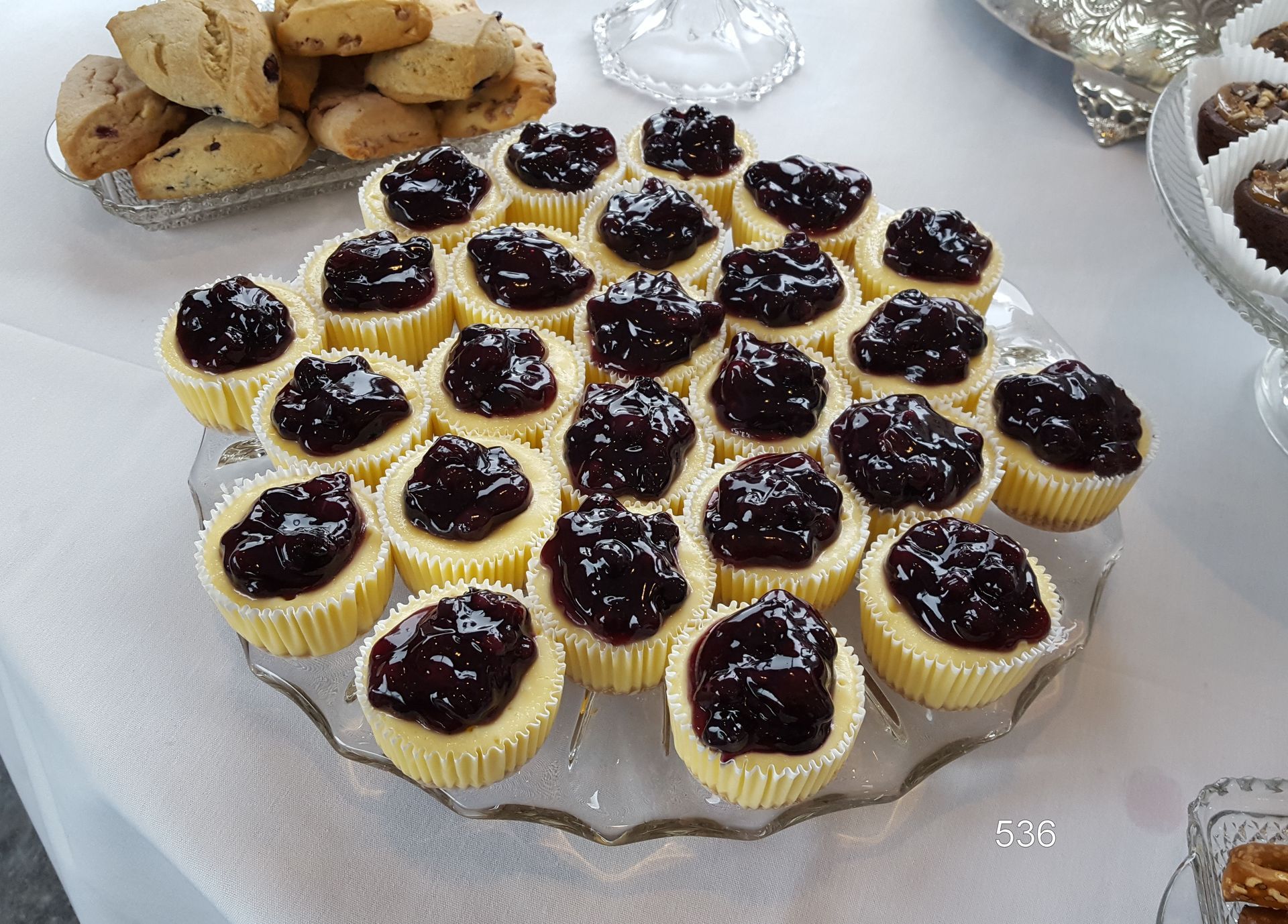 Tray of mini cheesecakes topped with dark berry jam, with cookies and a dessert bowl in the background