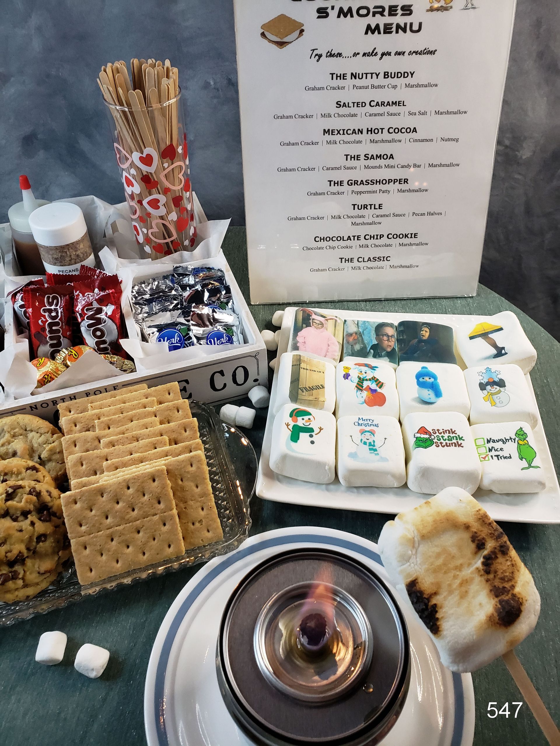 Baked goods and packaged snacks arranged on a table beside a “S’mores Menu” sign and marshmallows.