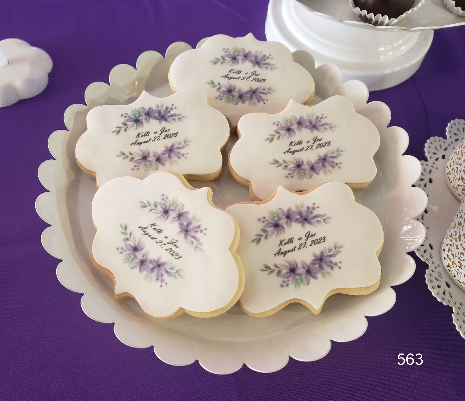 White flower-shaped cookies with purple lavender designs on a tray over a purple cloth.
