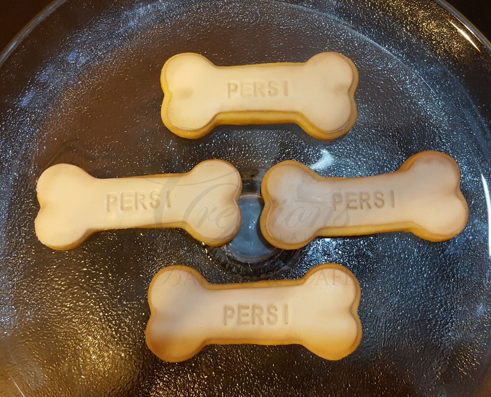 Four bone-shaped dog biscuits on a dark speckled pan, one pale and three tan, each embossed with “PERSI”.