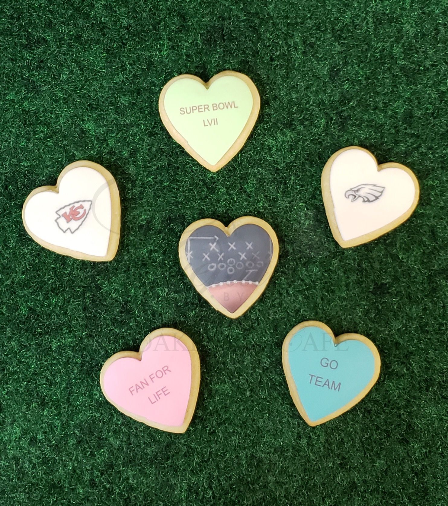 Five heart-shaped decorated cookies on green grass, in white, gold, pink, blue, and dark colors.