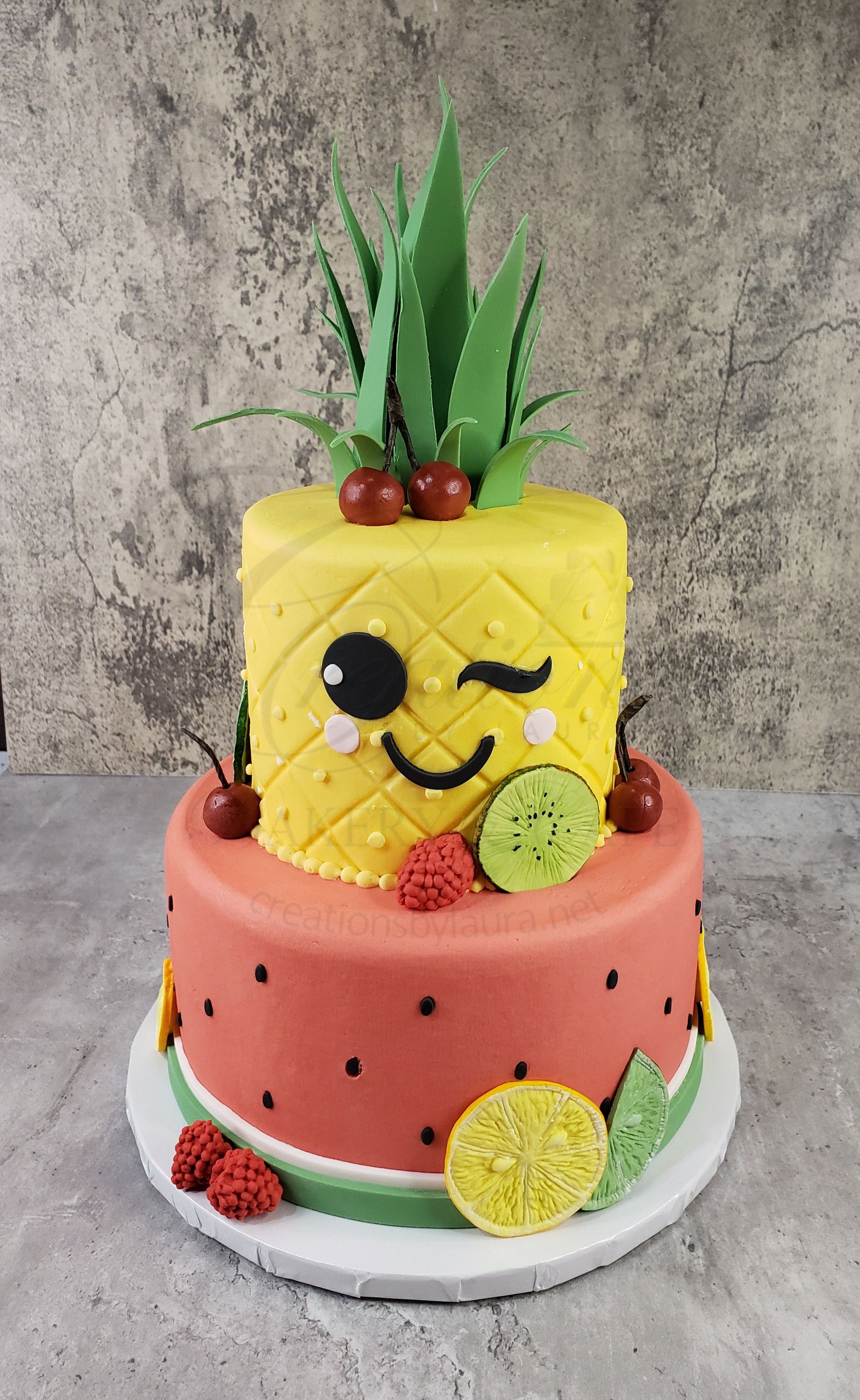 Two-tier pineapple and watermelon-themed cake with fruit decorations and a winking face