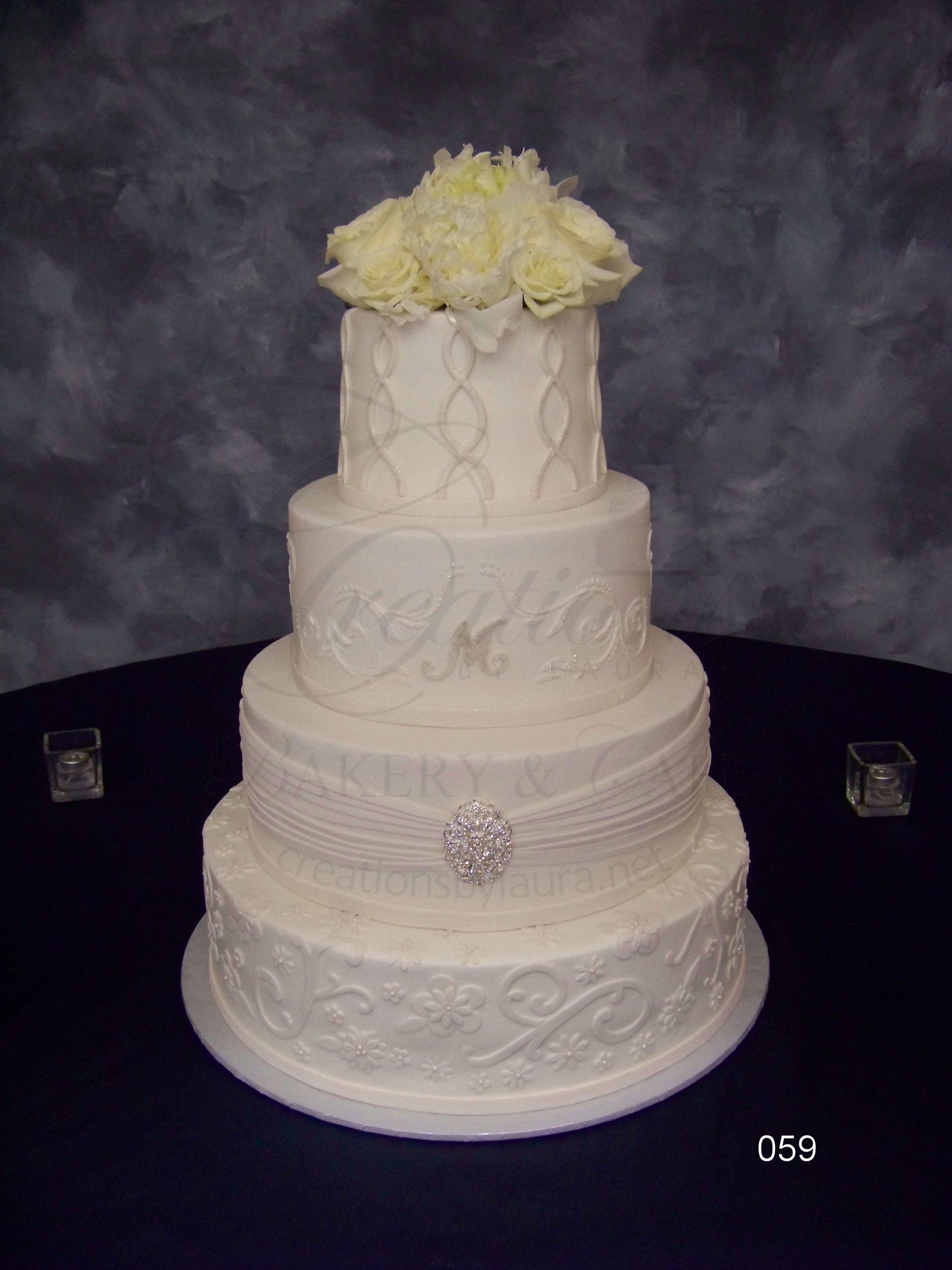 Four-tier white wedding cake with lace details, white flowers on top, and a jeweled brooch accent