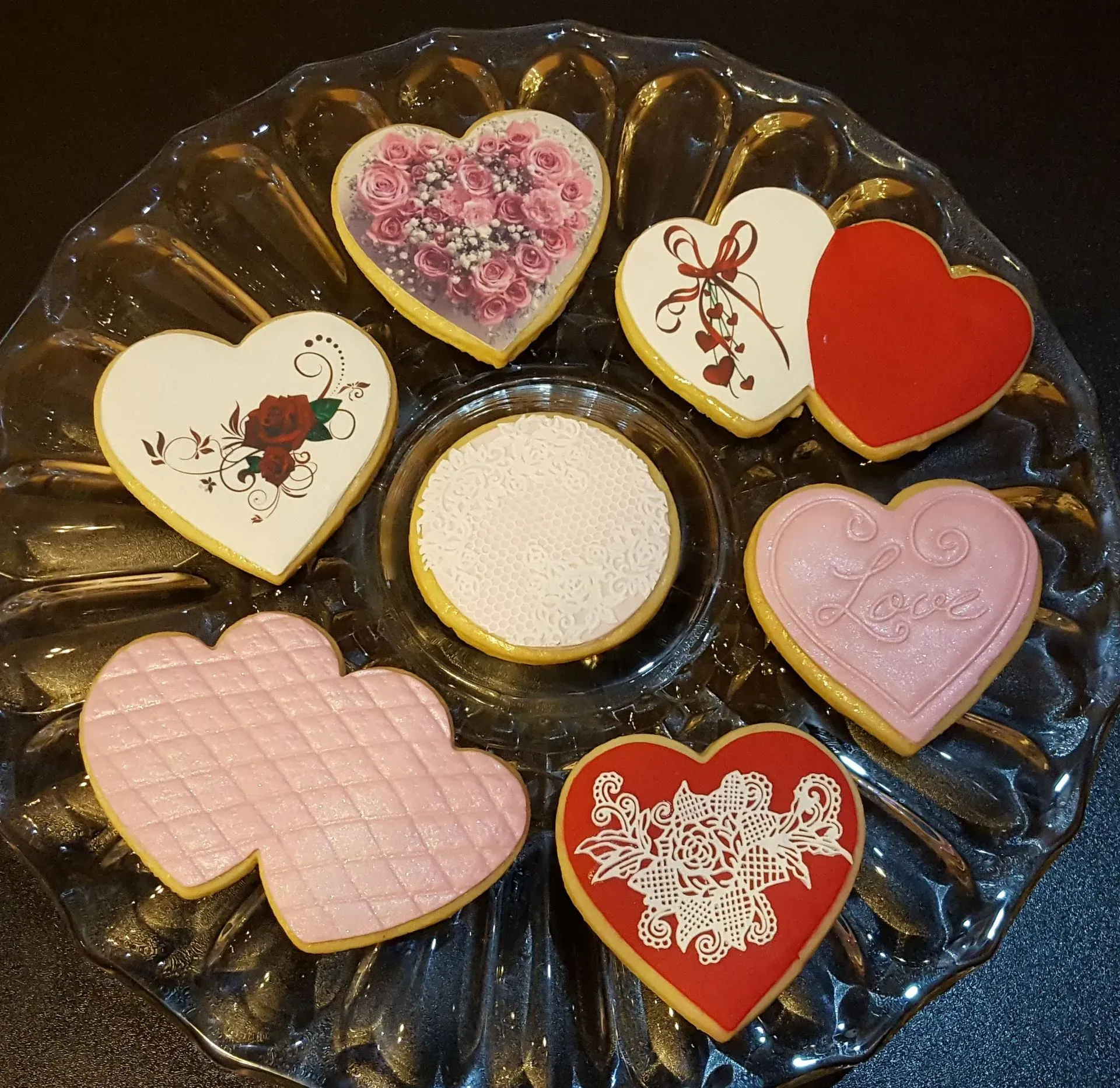 A glass plate holds eight heart-shaped cookies with various intricate white, pink, and red icing designs.