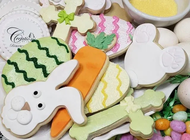 A platter of decorated Easter sugar cookies shaped like bunnies, carrots, crosses, and eggs, alongside pastel candies.