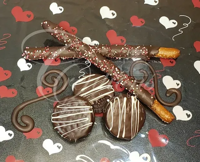 Chocolate-dipped pretzel sticks and round cookies with white drizzle on a heart-patterned background