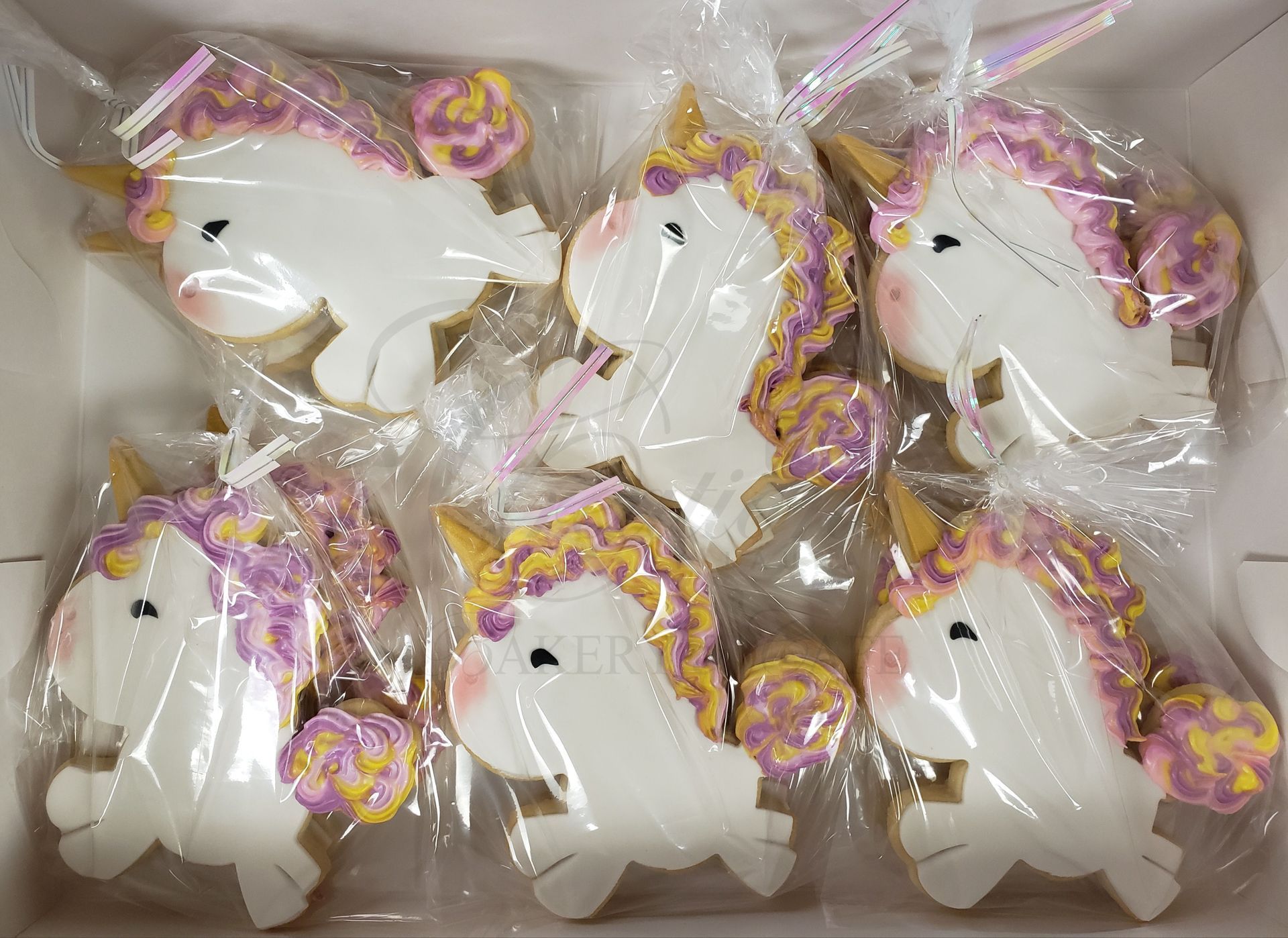 Six unicorn-shaped cookies in clear plastic wrappers, white with pink and yellow icing accents.