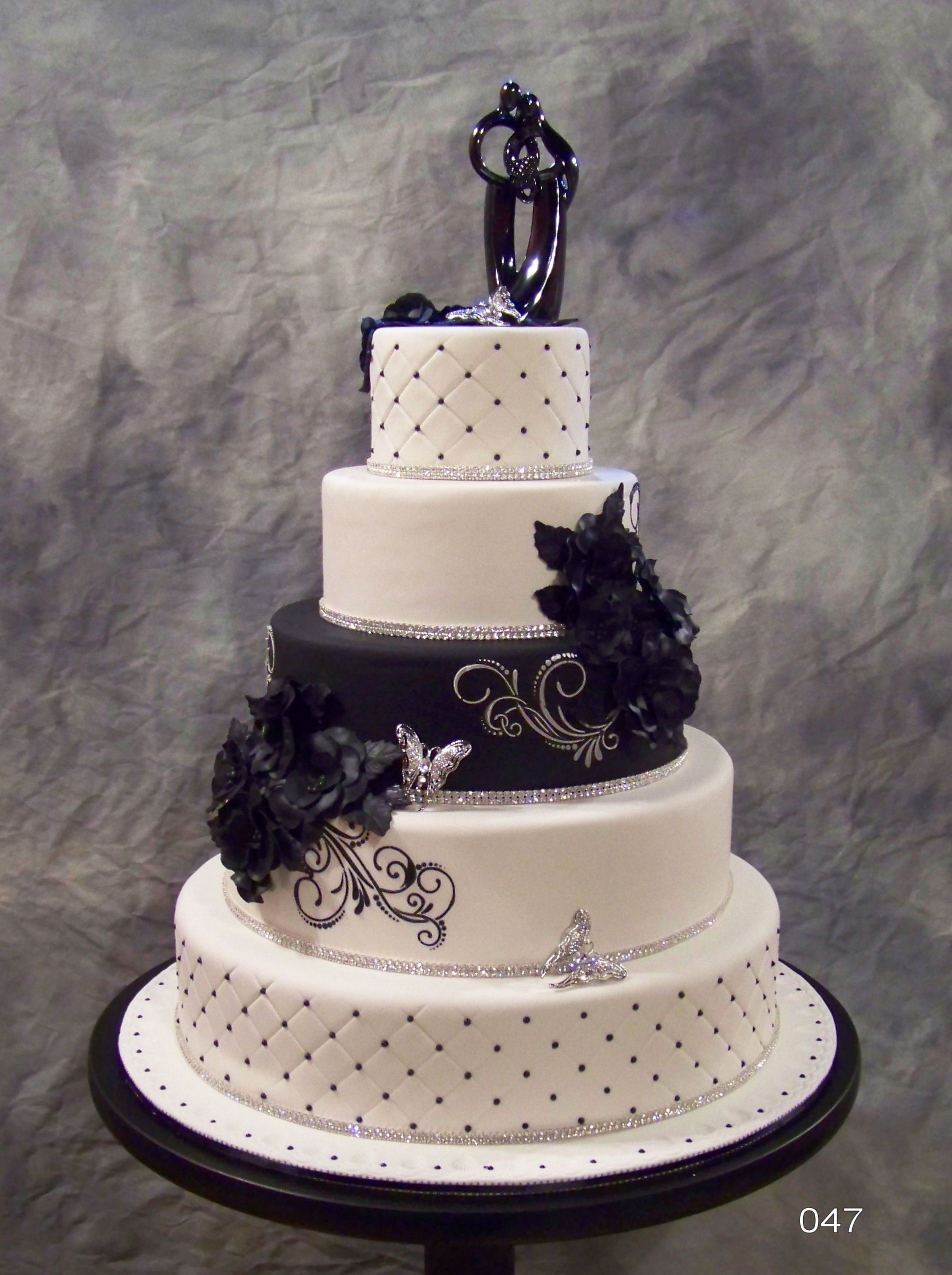 A five-tier wedding cake in black and white with quilted patterns, decorative swirls, dark floral accents, and a topper.