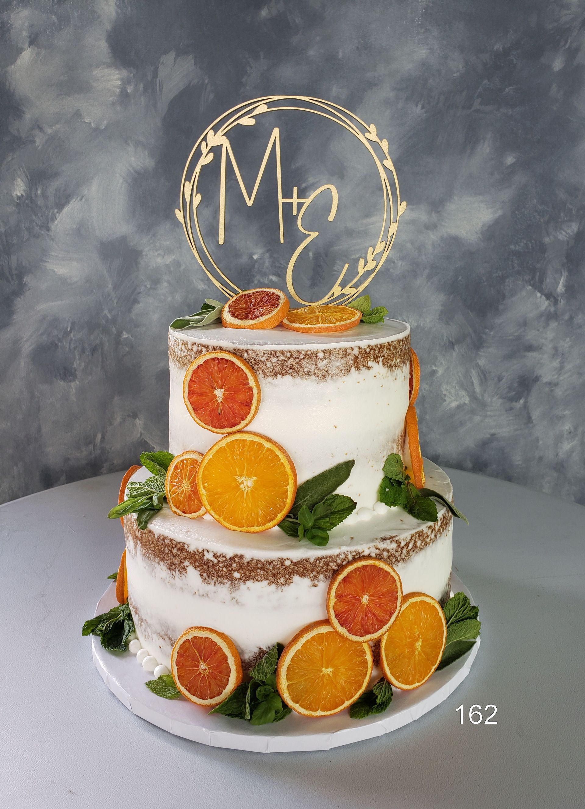 A two-tiered, white-frosted cake decorated with fresh orange slices and mint leaves, topped with a gold 