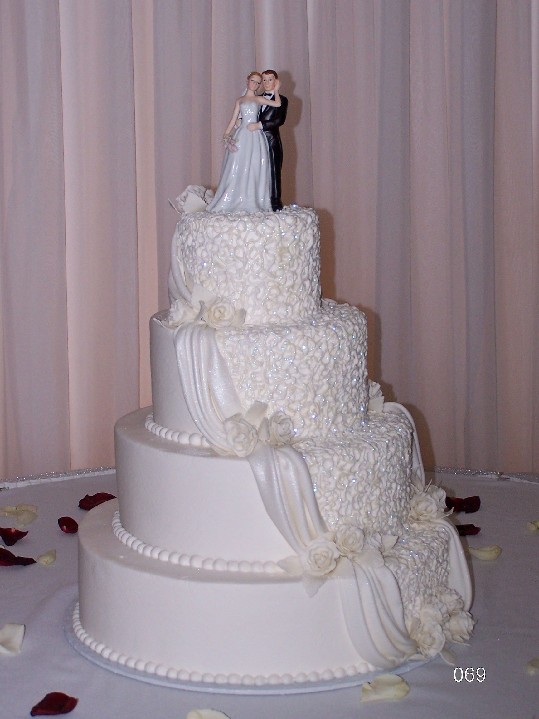 A four-tiered white wedding cake with textured icing, decorative fabric drapes, and a small bride and groom topper.