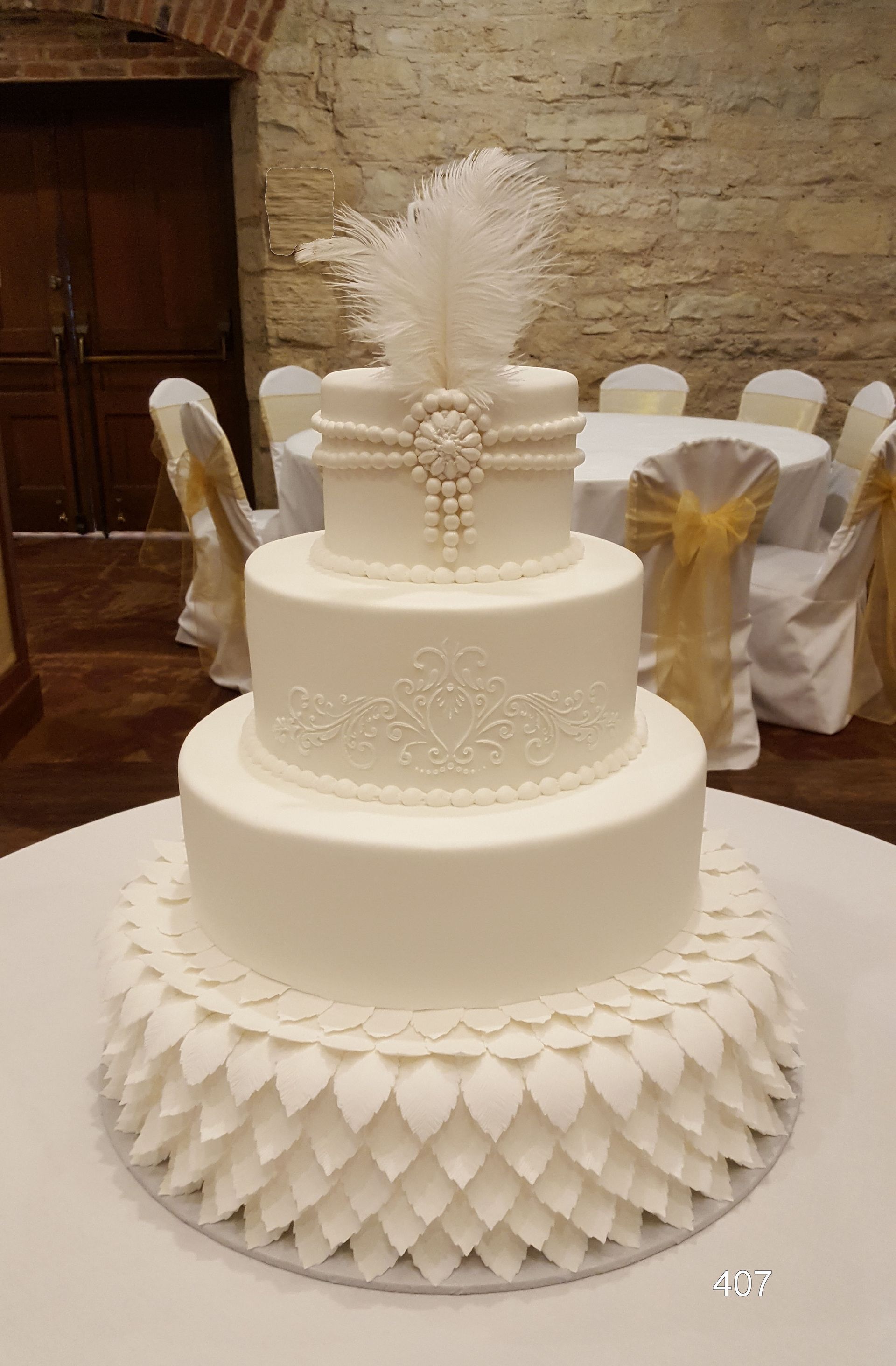 A four-tier white wedding cake with a feathered topper and pearl decorations, set on a table in a banquet hall.