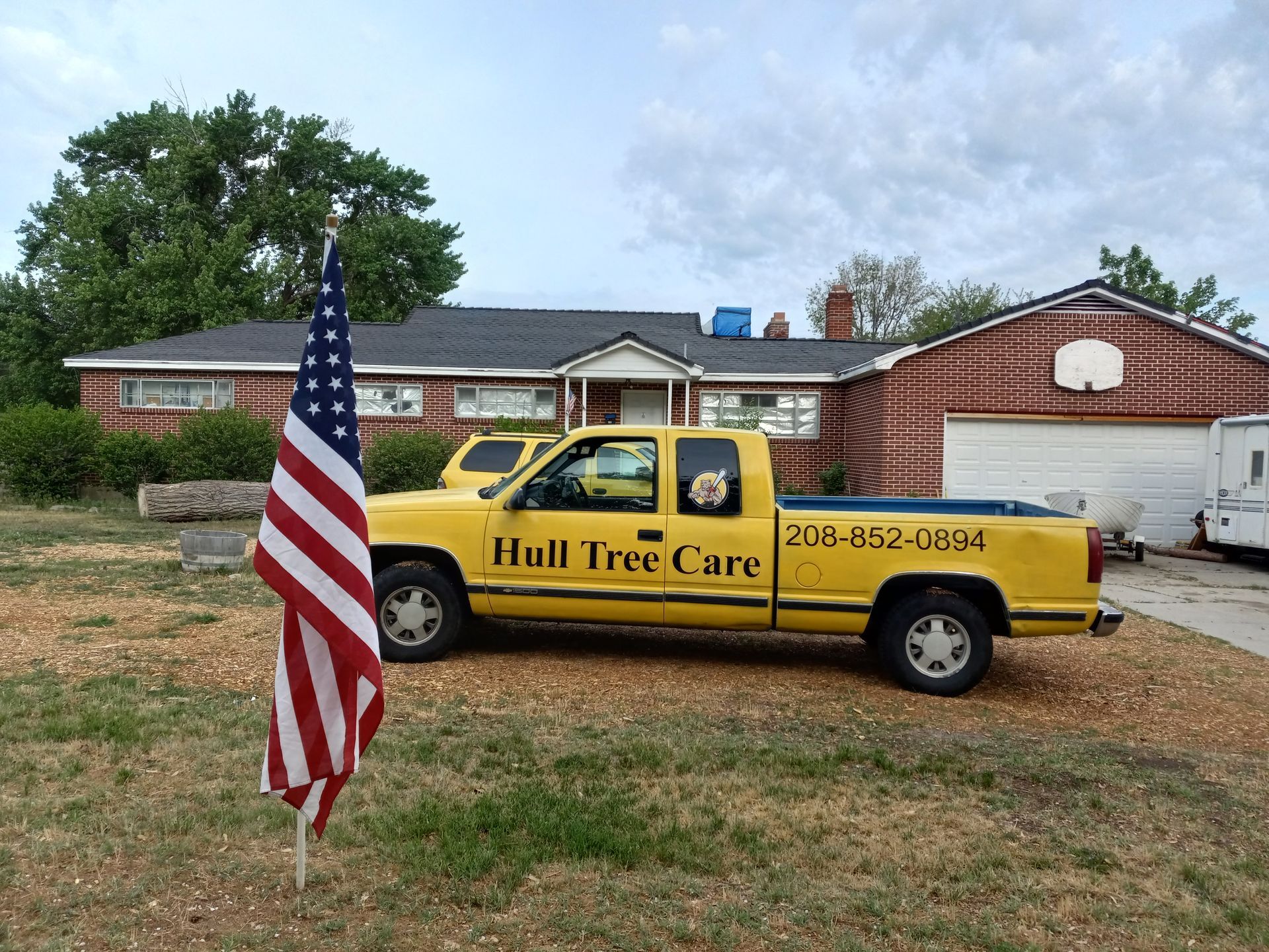 Yellow Hull Tree Care truck parked on grass beside a U.S. flag in front of a house and garage.