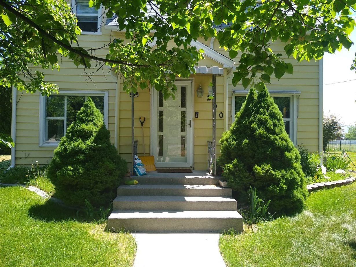 Yellow house with front steps, white trim, and neatly trimmed shrubs under leafy trees.