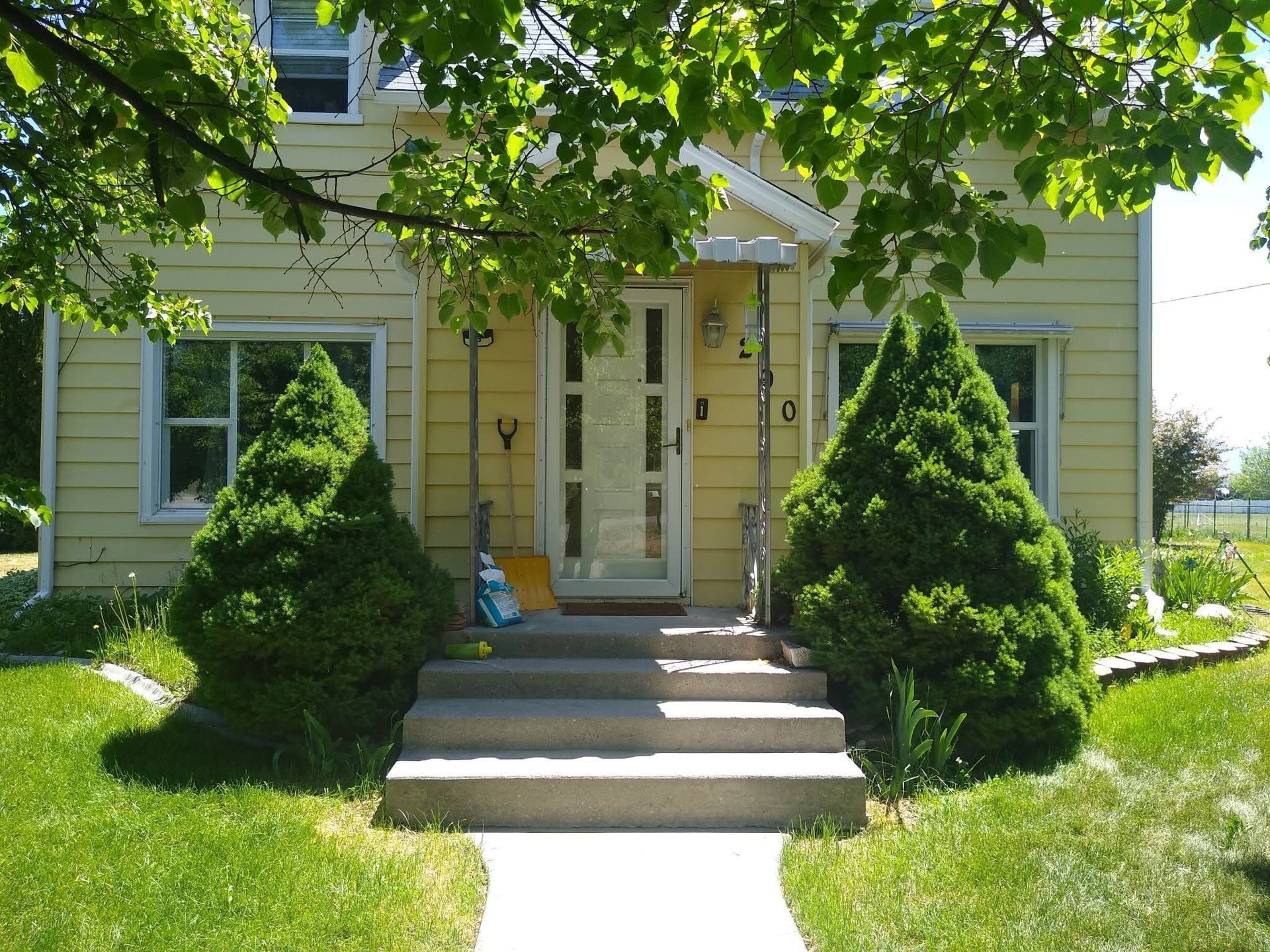 Yellow house with front steps, white trim, and neatly trimmed shrubs under leafy trees.