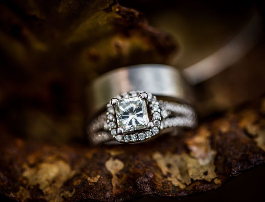 Wedding rings, diamond halo engagement ring in focus, silver band behind, on a textured brown surface.
