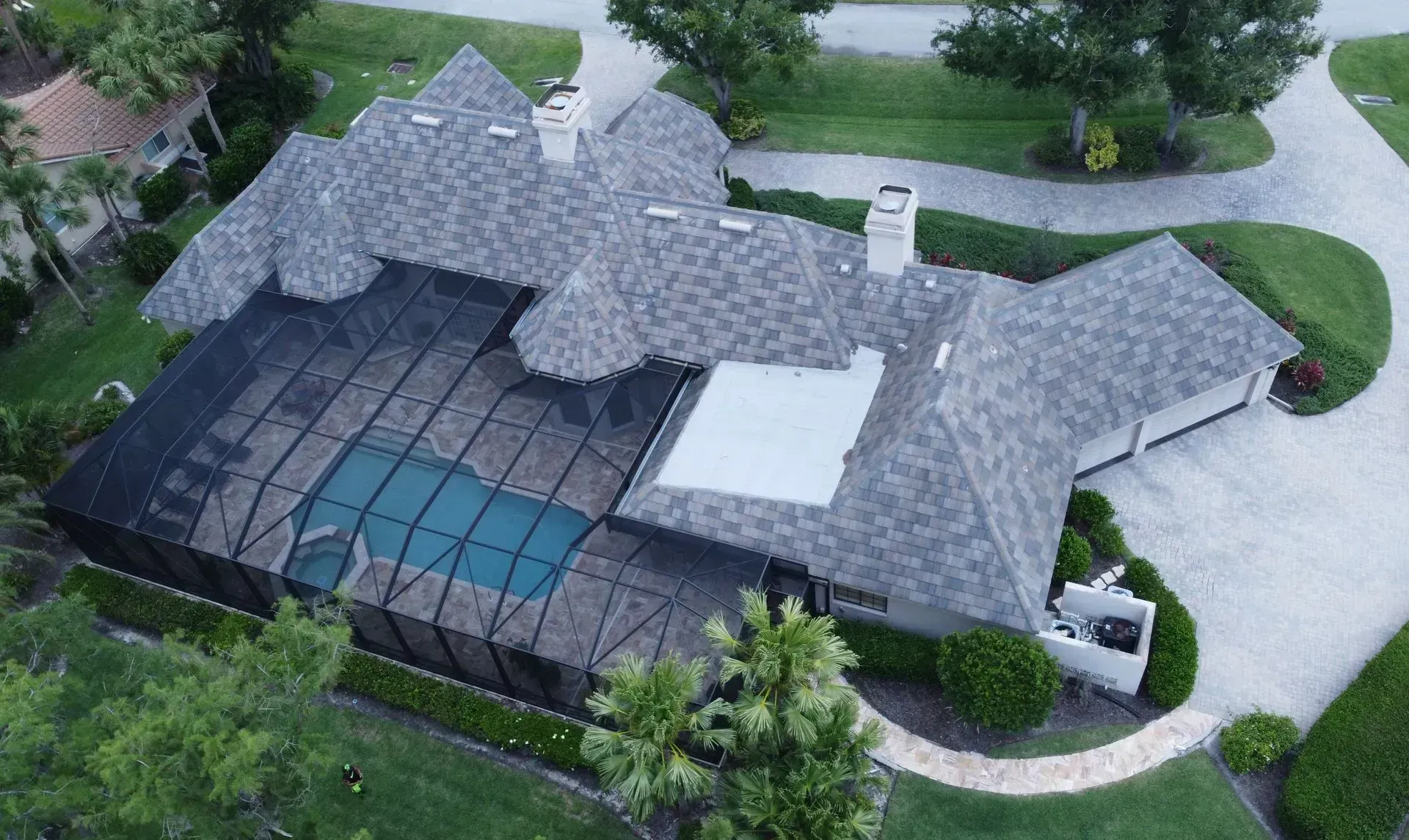 Aerial view of a house with a pool enclosed by a screen, gray roof, and a driveway.